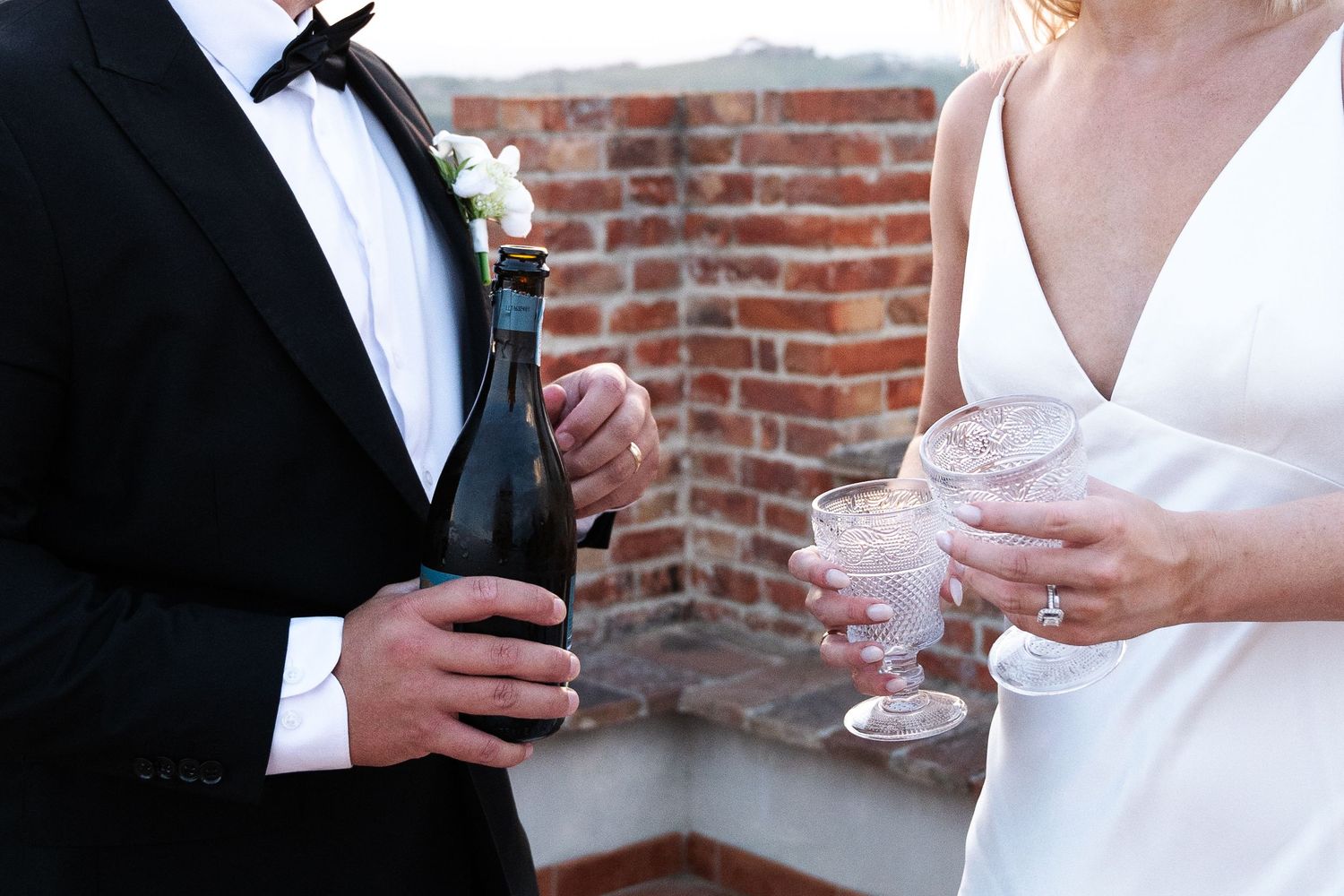 Bride and groom toasting with champagne glasses near a rustic brick wall at their wedding.