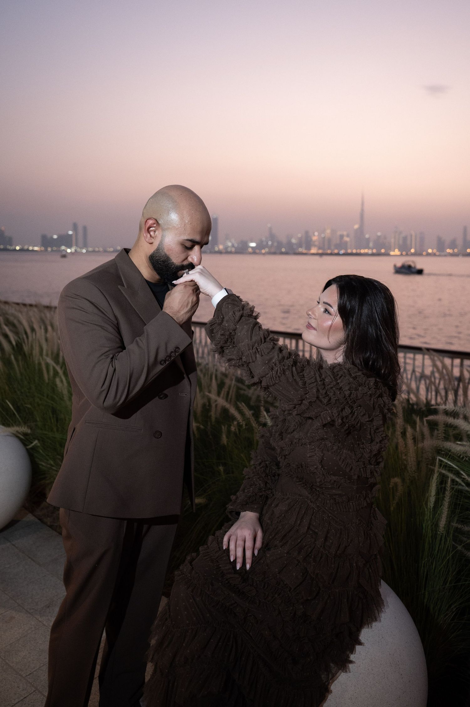 A couple shares a romantic moment at sunset with a city skyline backdrop, man kissing womans hand.
