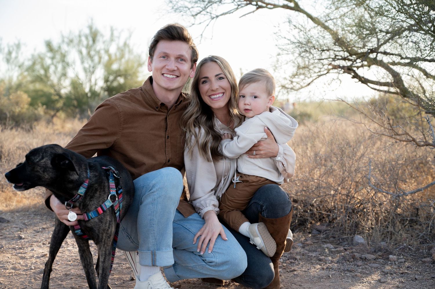 A smiling young family with a black dog poses outdoors in a desert landscape for a casual portrait photo.