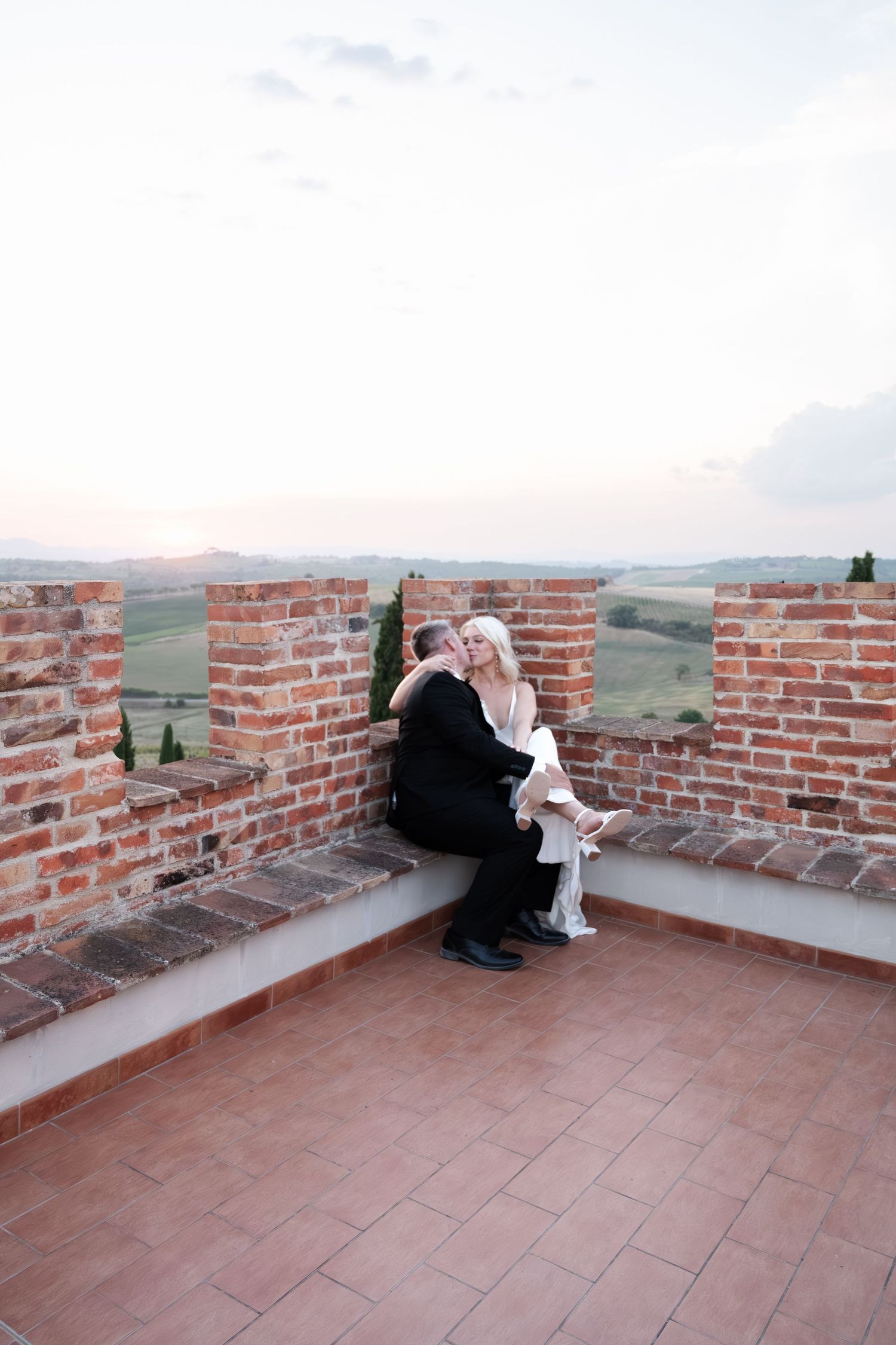 Newlyweds embrace on a rustic brick terrace overlooking rolling Tuscan hills at sunset.