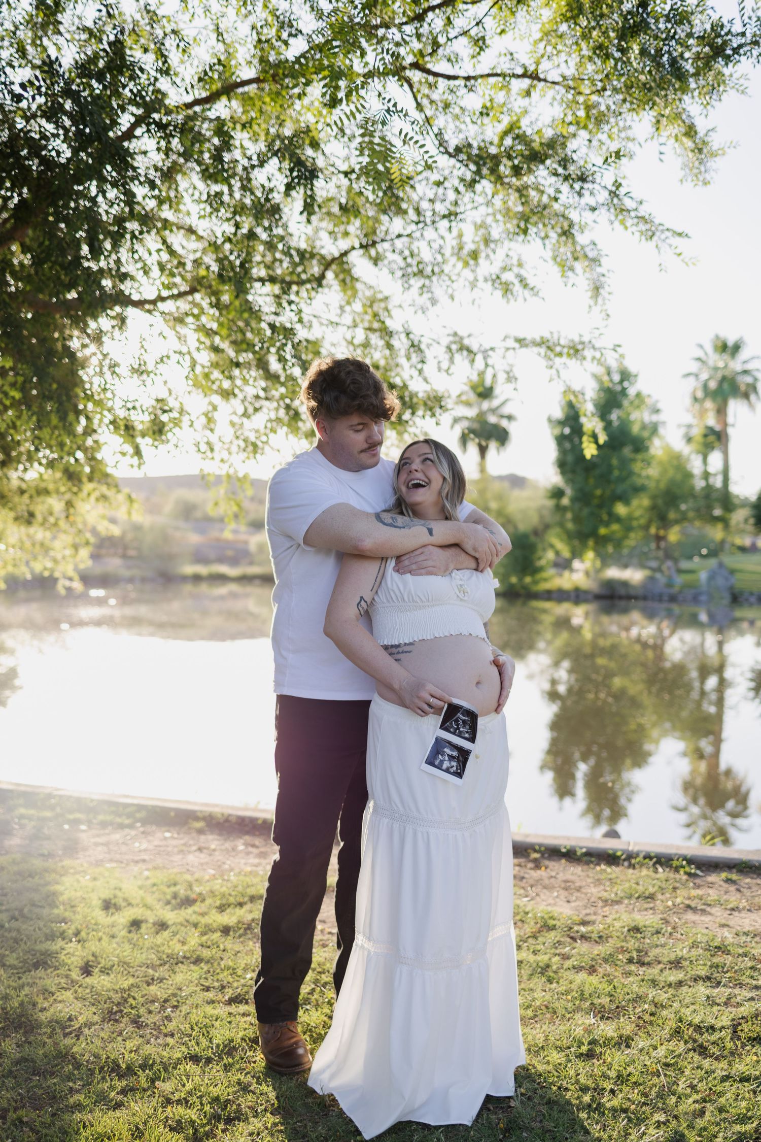 Laughing pregnant woman and partner hold ultrasound photo by sunlit lake under willow tree in phoenix arizona