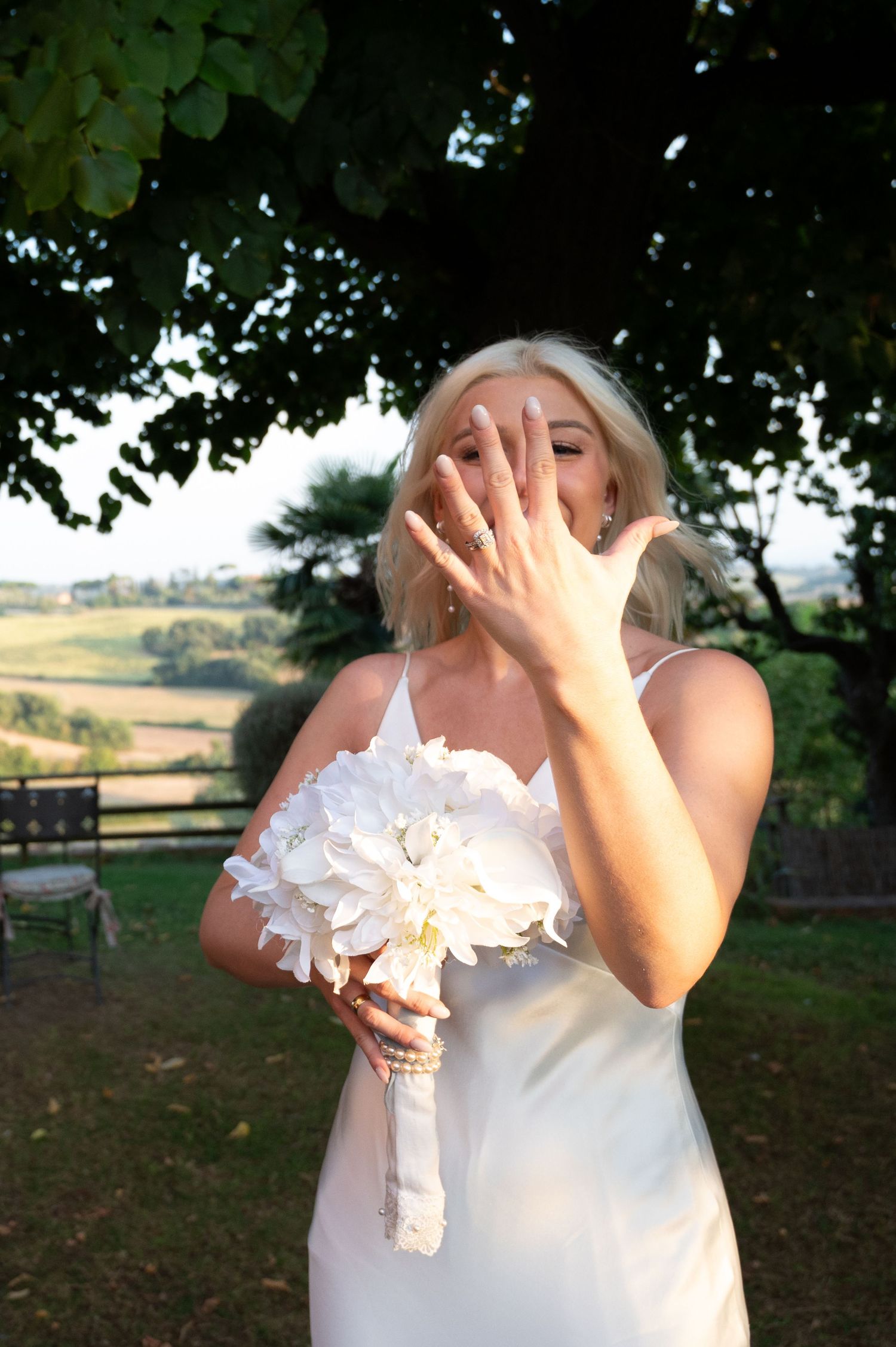 Joyful bride laughs and shows off her wedding ring while holding a white bouquet beneath a leafy tree in Tuscany Italy.