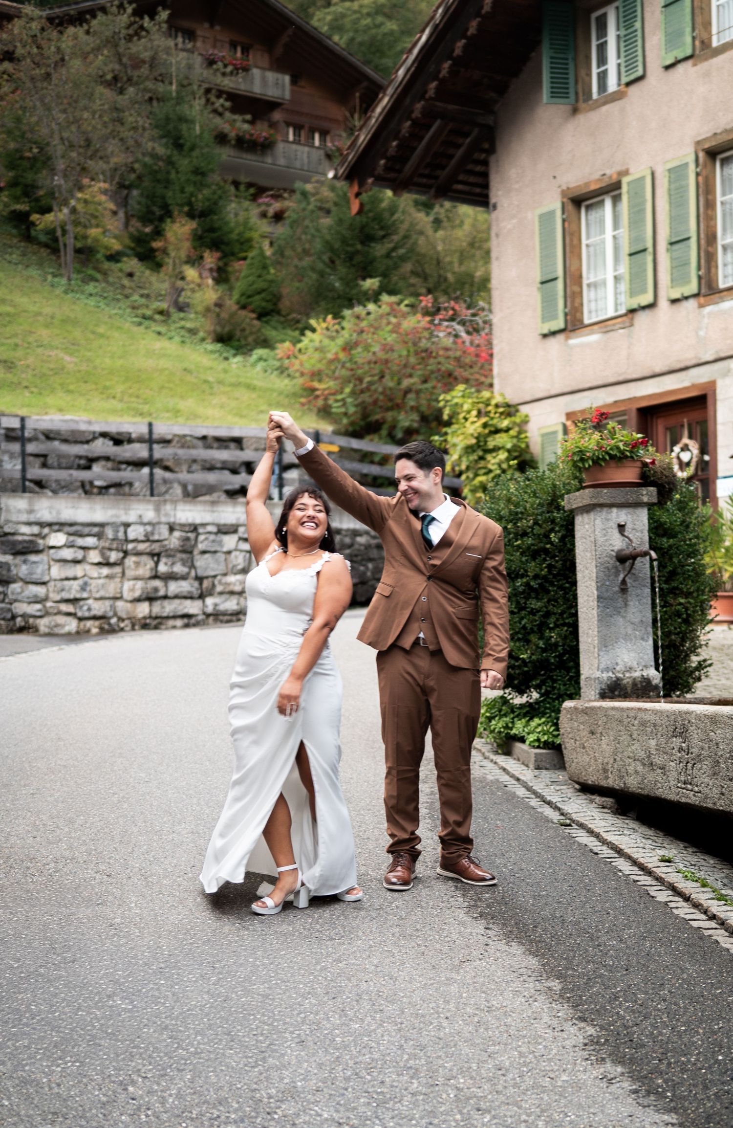 Joyful bride twirling in white gown as groom spins her on charming Swiss alpine village cobblestone street in Lauterbrunnen Switzerland.