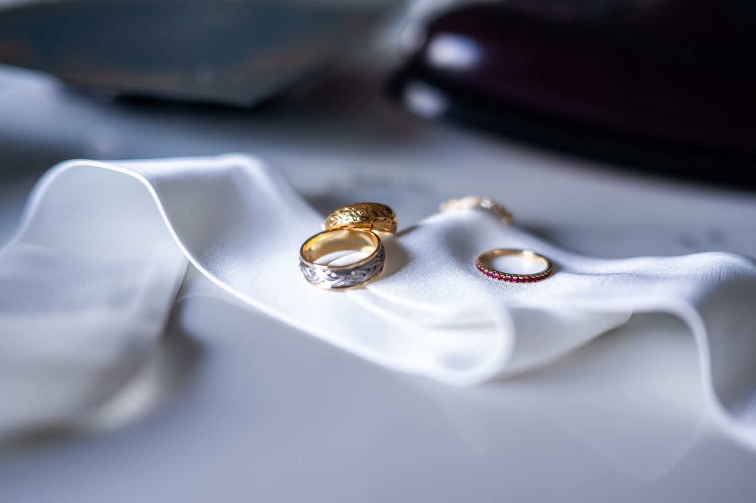 Gold wedding bands and diamond ring arranged on white silk ribbon on white surface.