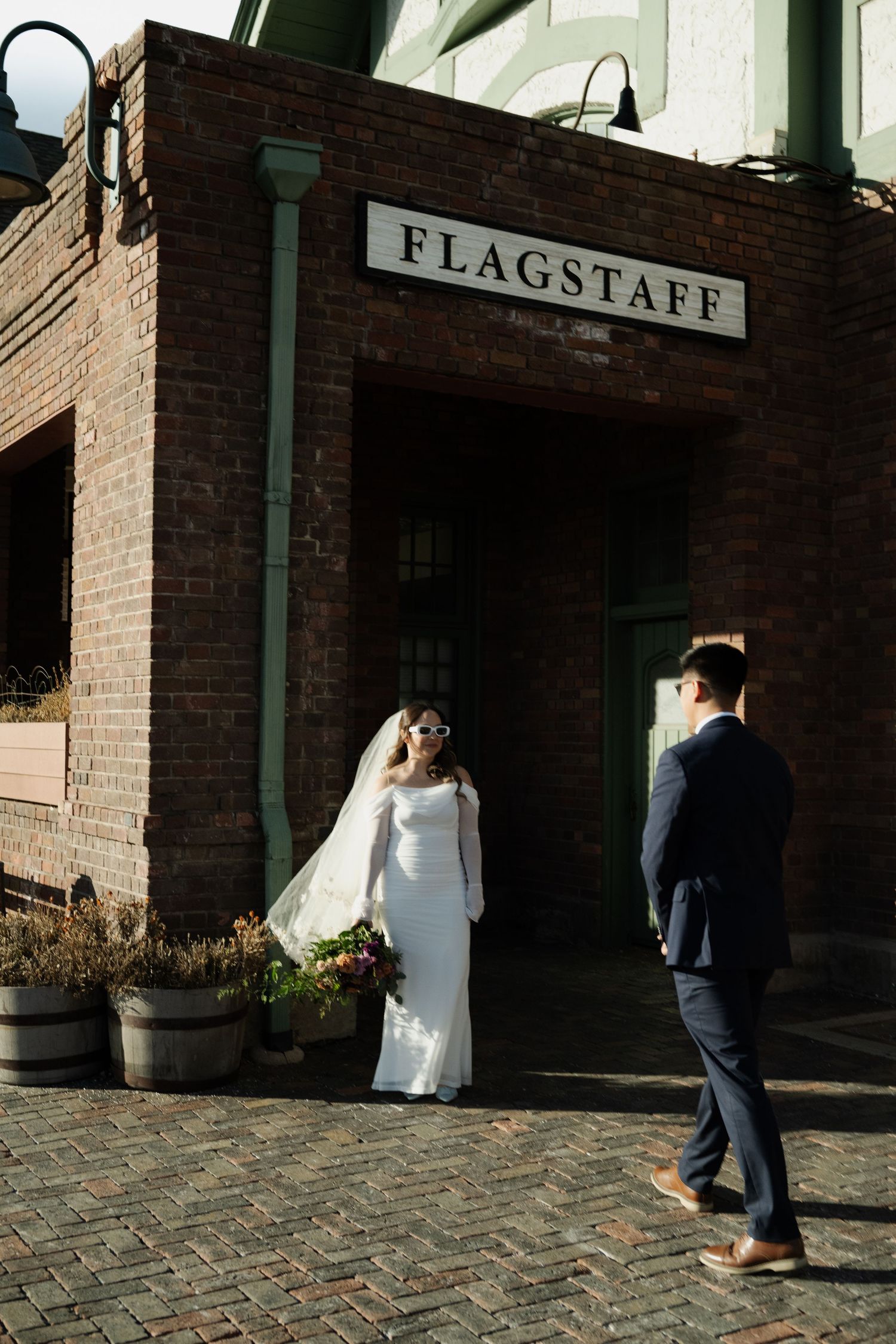 Bride in white gown with veil poses before Flagstaff brick building as groom approaches in Flagstaff Arizona.