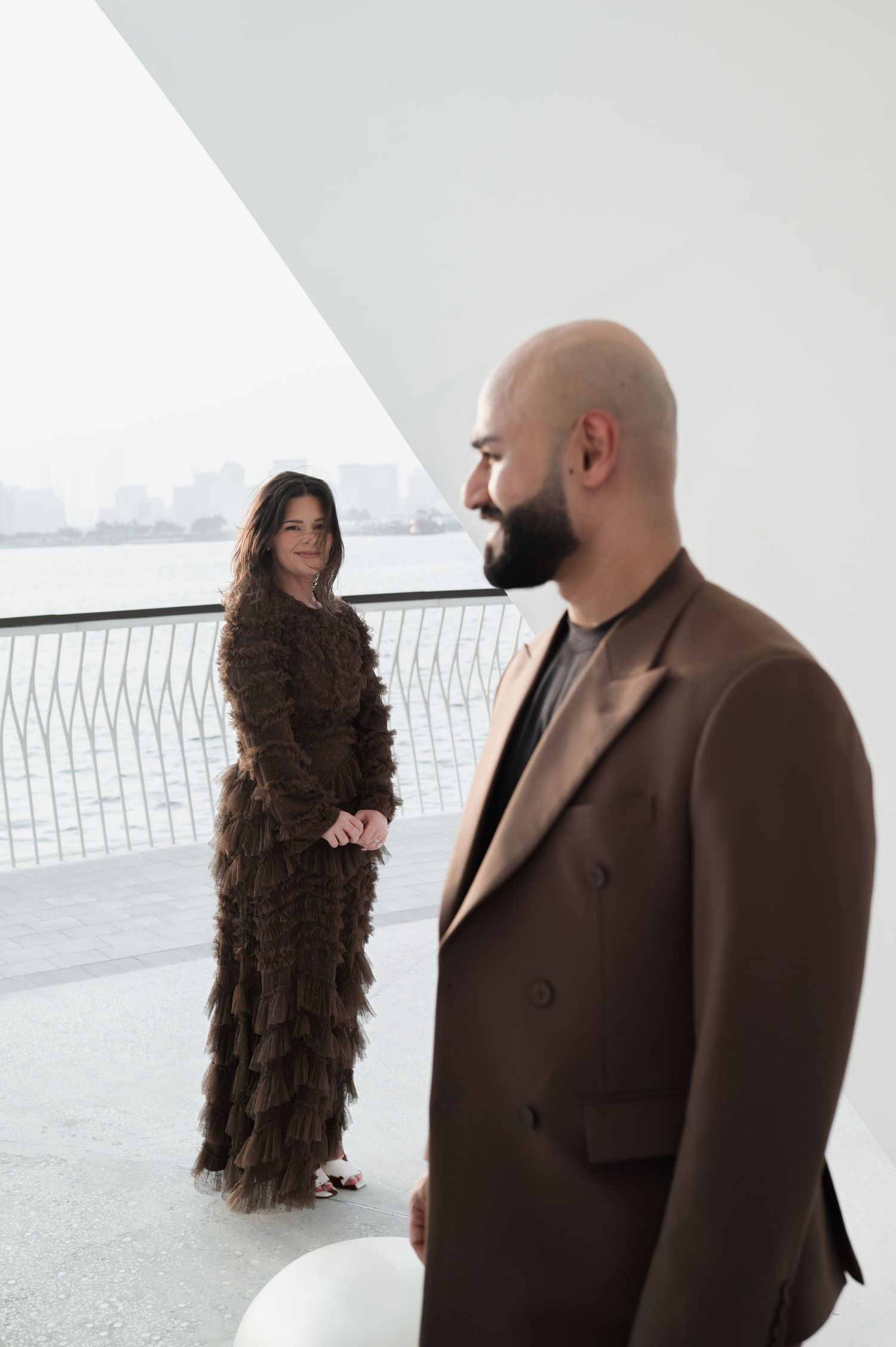 A bald bearded man in a brown suit stands in the foreground as a woman in a brown ruffled gown poses behind him.
