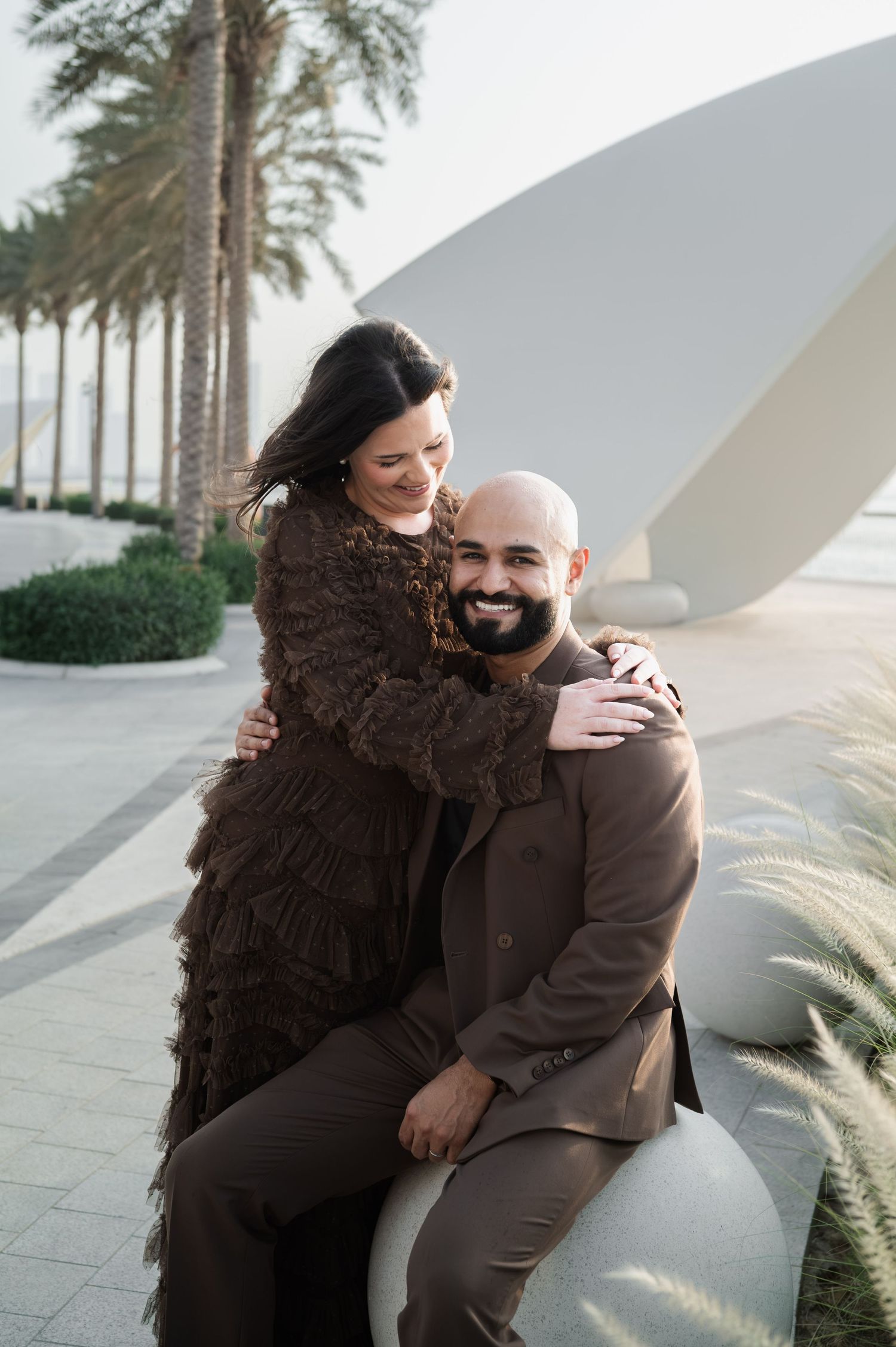 A smiling couple in matching brown outfits poses outdoors near palm trees, the woman embracing the seated man affectionately.