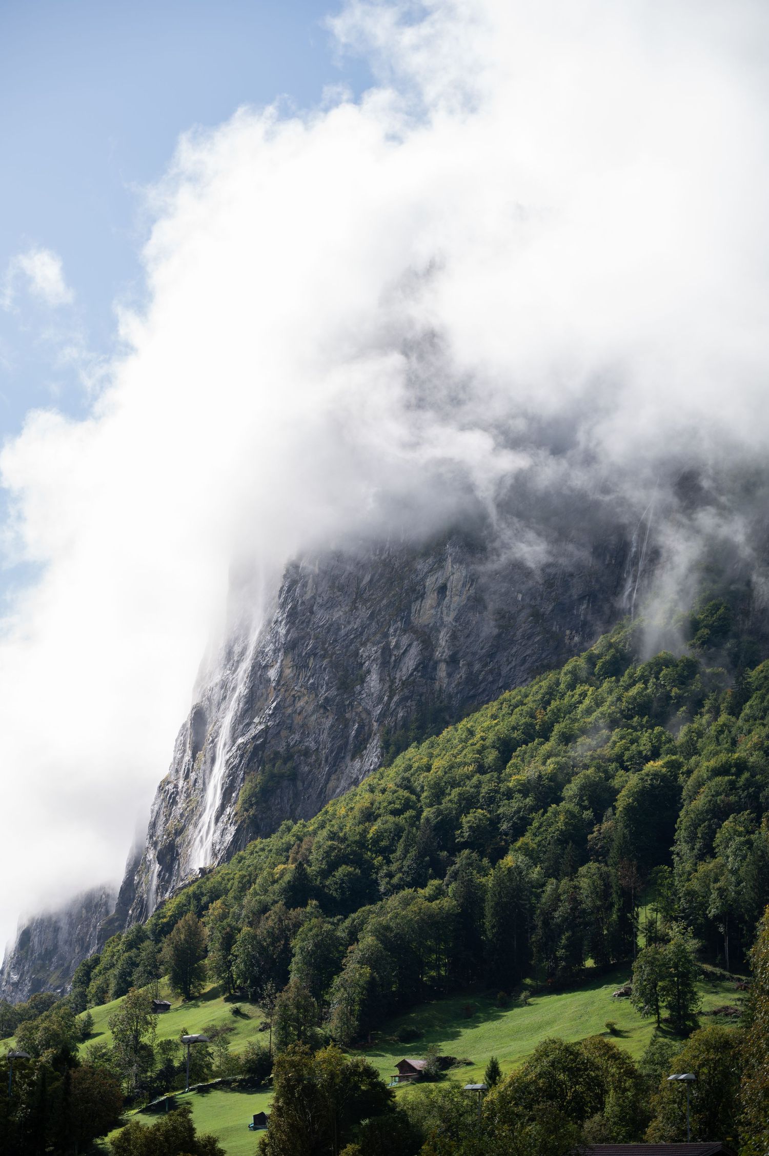 Dramatic Swiss mountain cliff shrouded in clouds above lush green forest and alpine meadow valley in Switzerland.