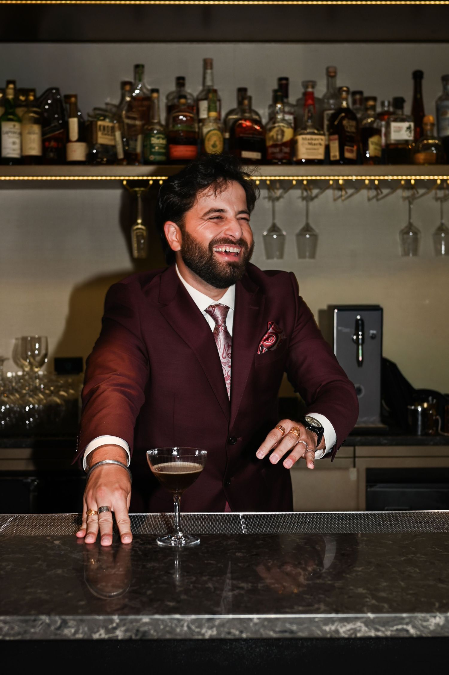 Smiling groom in burgundy suit leans on marble bar with a chocolate cocktail at wedding reception at The Pendry Hotel in Chicago Illinois.