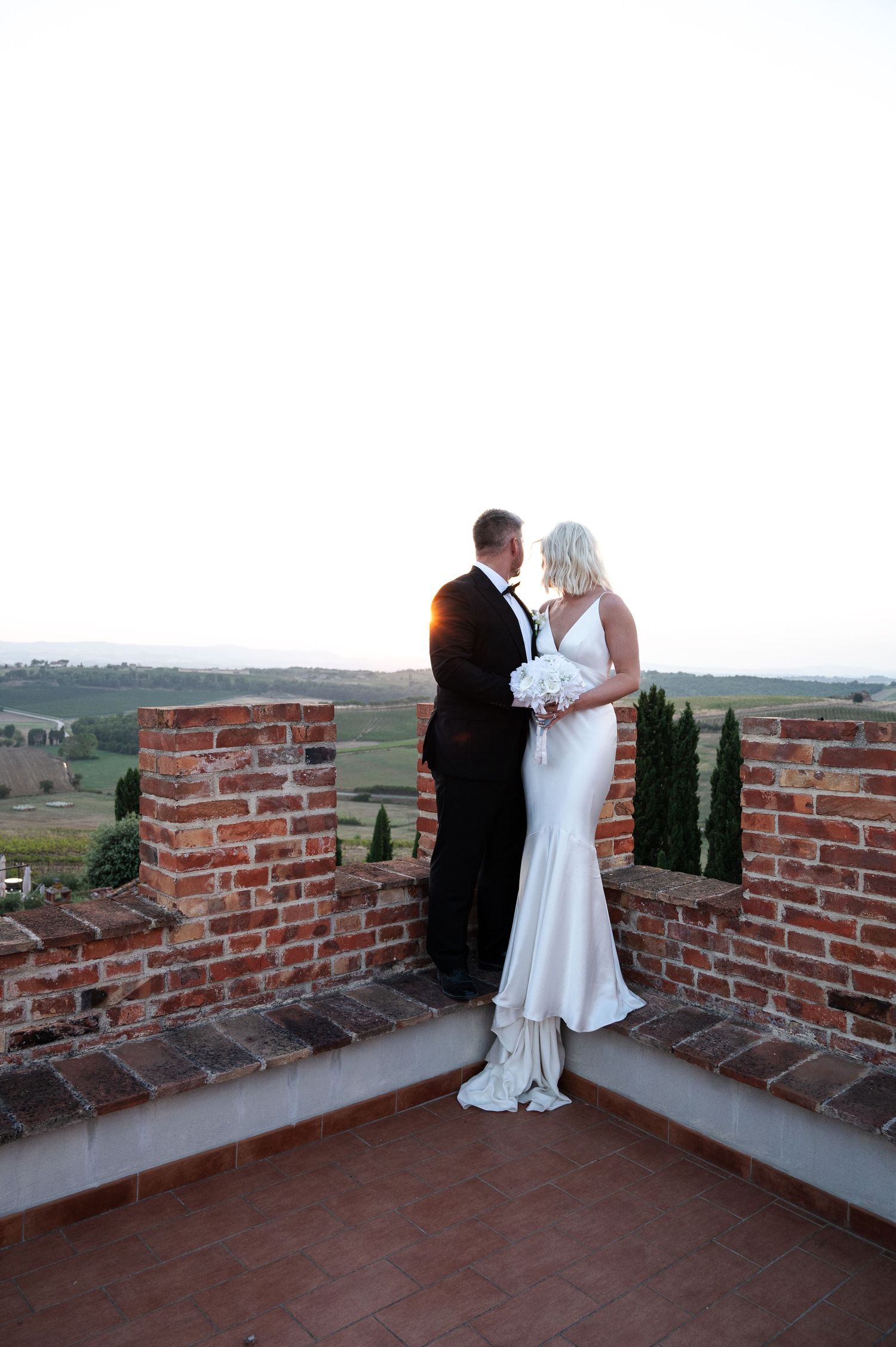 Newlywed couple sharing romantic moment on rustic brick terrace overlooking Tuscan countryside at sunset in Tuscany Italy.