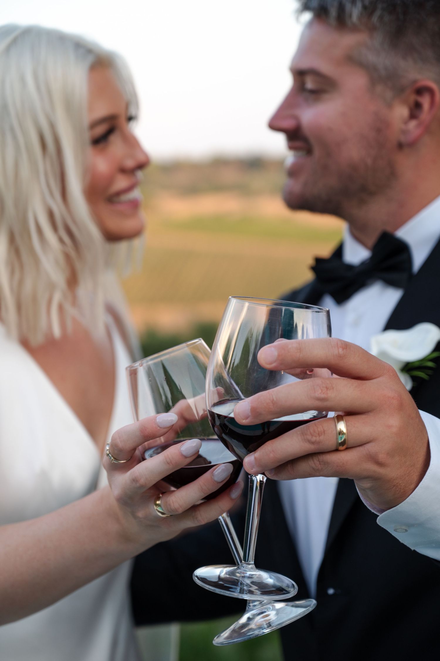 Bride and groom clinking red wine glasses during outdoor wedding celebration with scenic vineyard backdrop in Tuscany Italy.