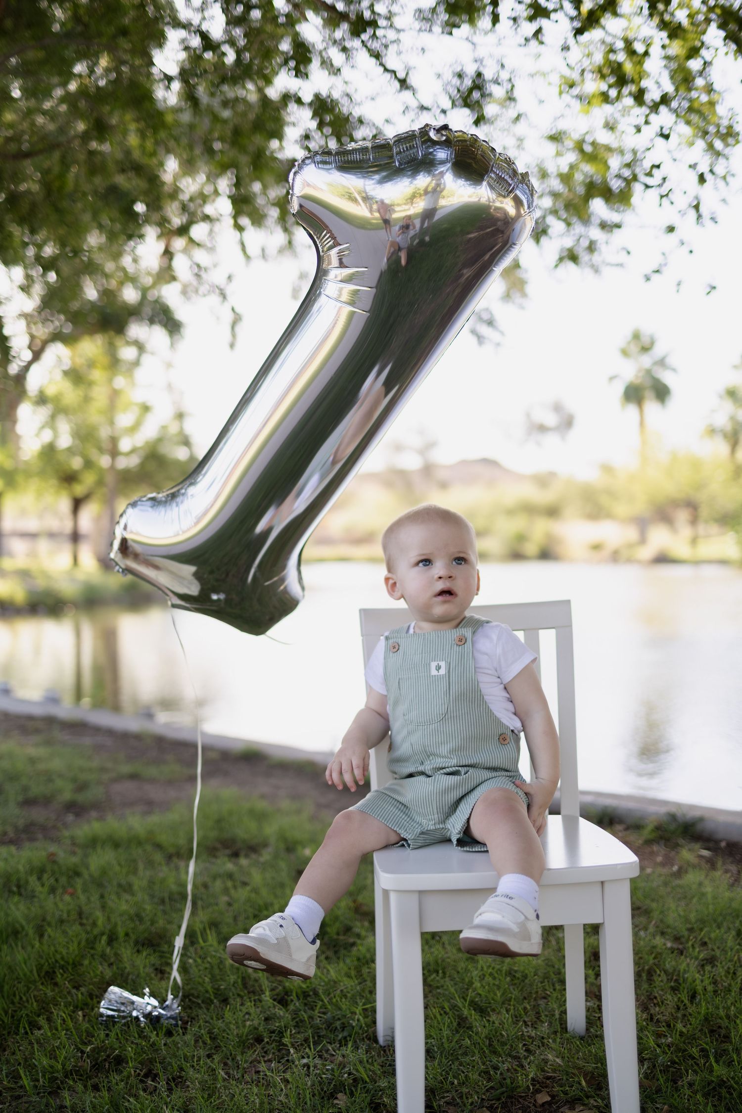 Baby in green overalls sits on white chair with silver number 1 balloon at outdoor first birthday celebration.