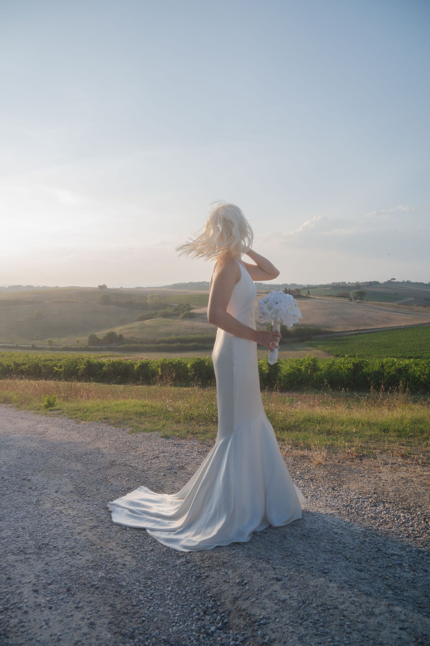 Bride in elegant white mermaid gown holding bouquet on scenic Tuscany countryside road at golden hour in Tuscany Italy.