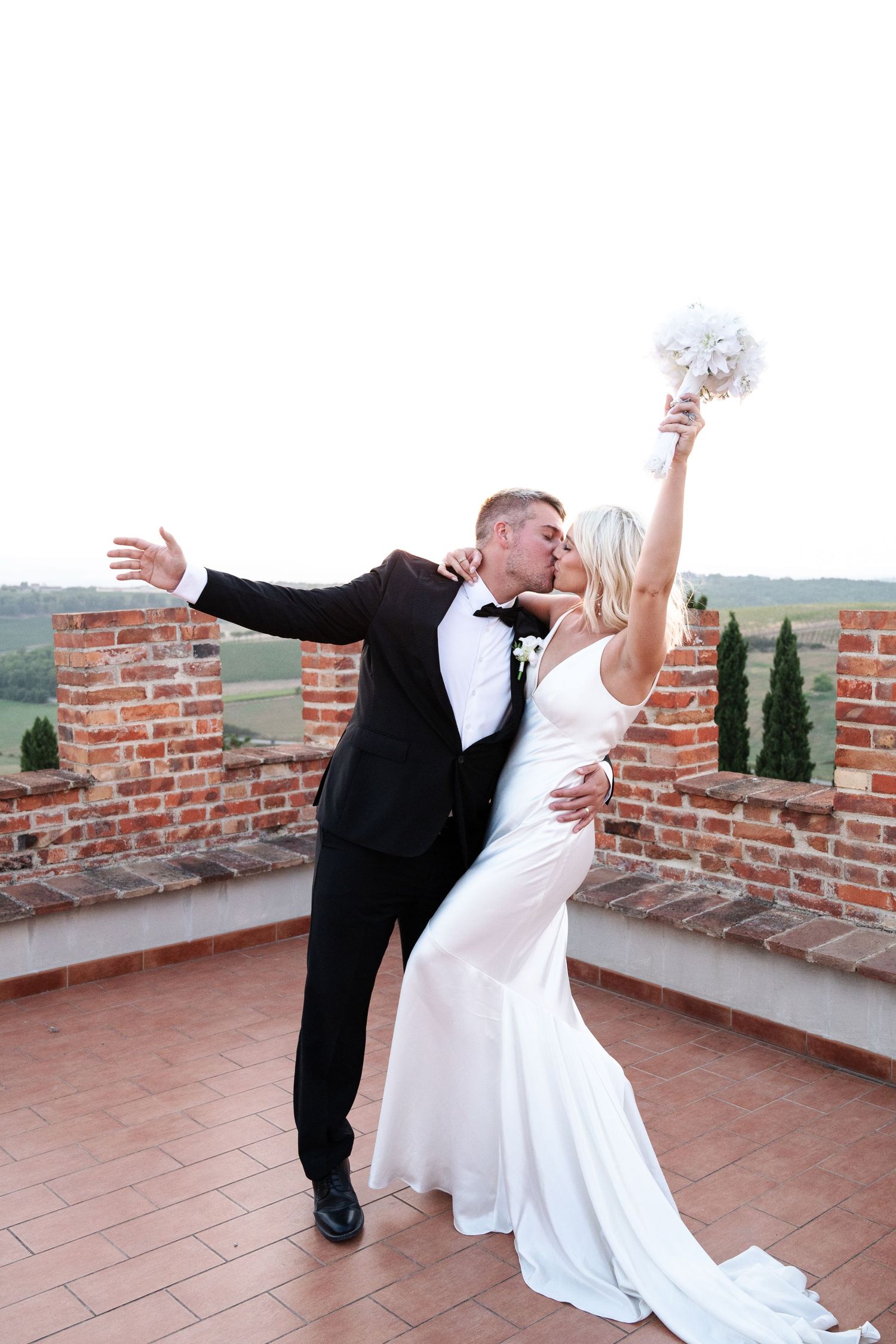 Groom dips bride for a kiss on a rooftop terrace as she raises her bouquet triumphantly with Tuscany views in Tuscany Italy.