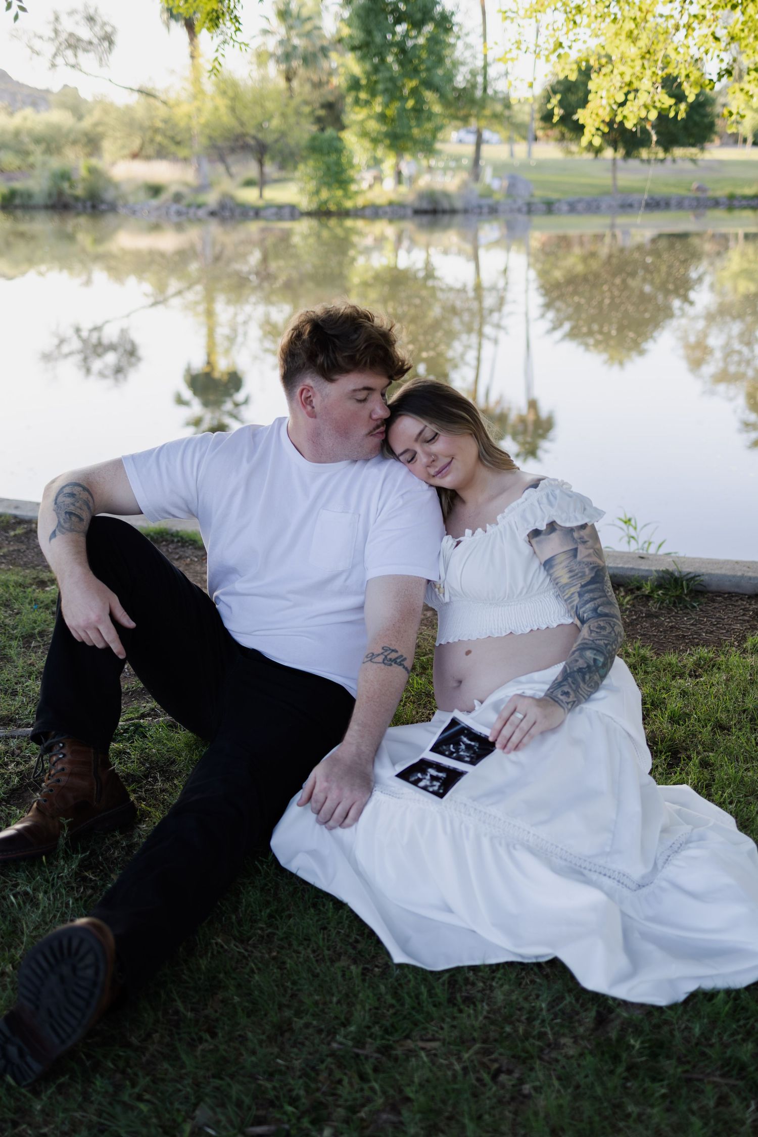 Couple sitting by a lake holding an ultrasound photo, sharing a tender pregnancy announcement moment.