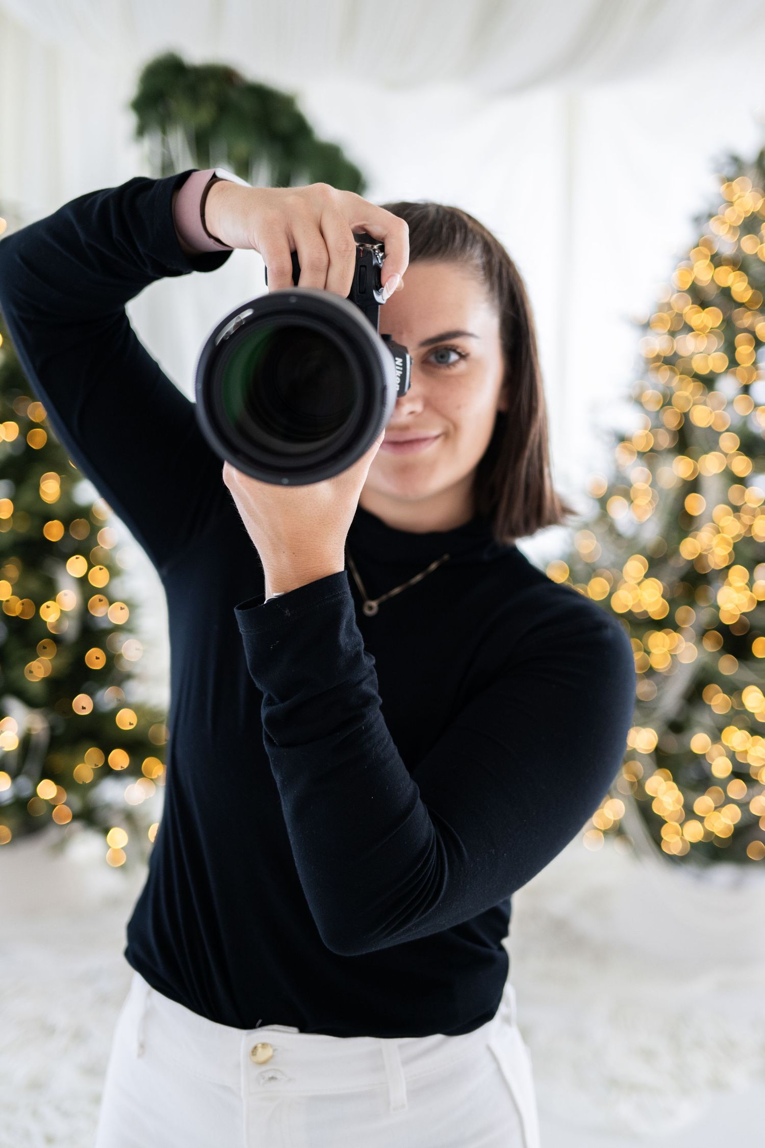Woman photographer smiling while holding a DSLR camera in front of decorated Christmas trees with golden bokeh lights.