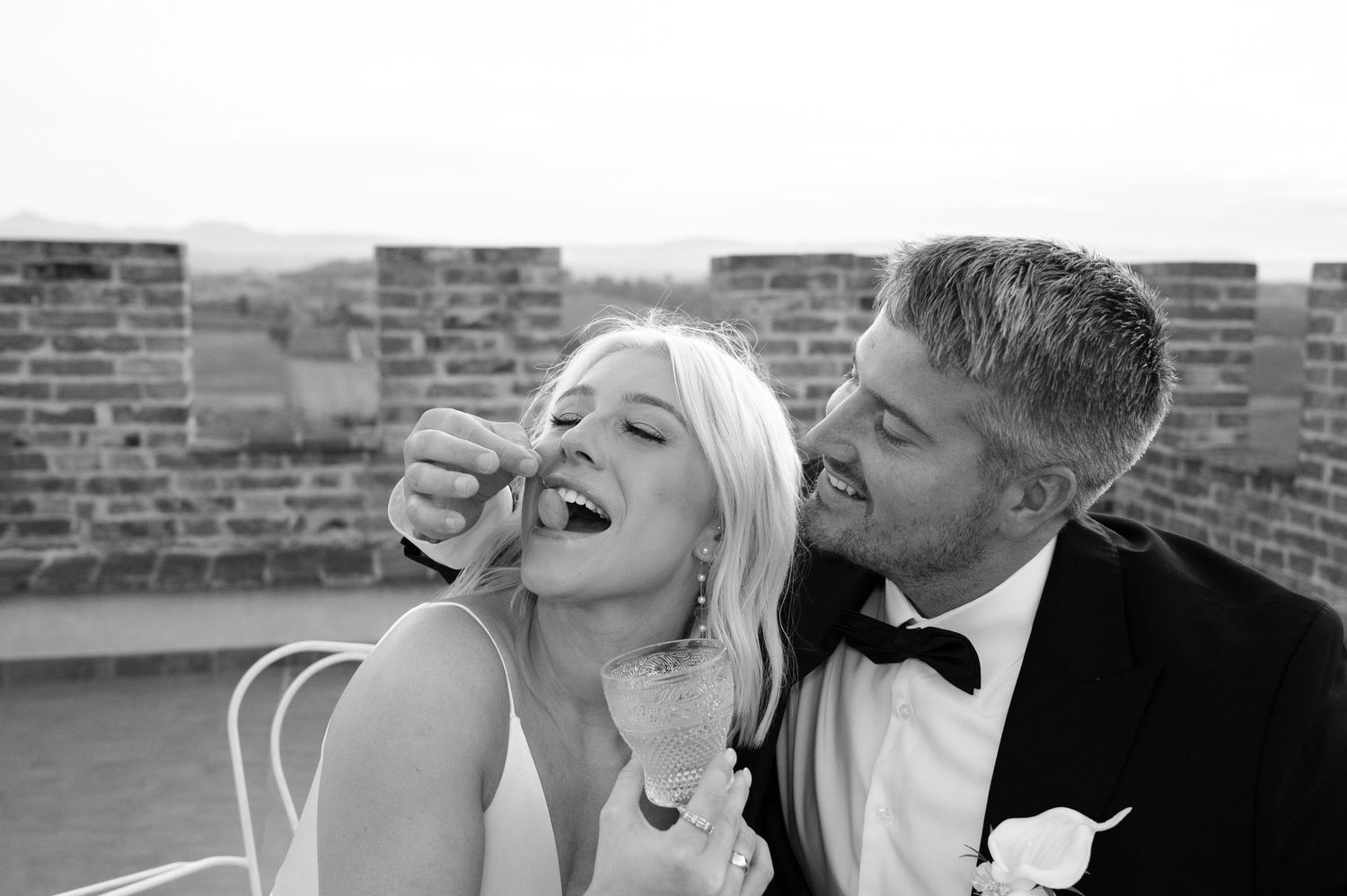 Black and white photo of a groom feeding his bride wedding cake, both smiling joyfully outdoors.