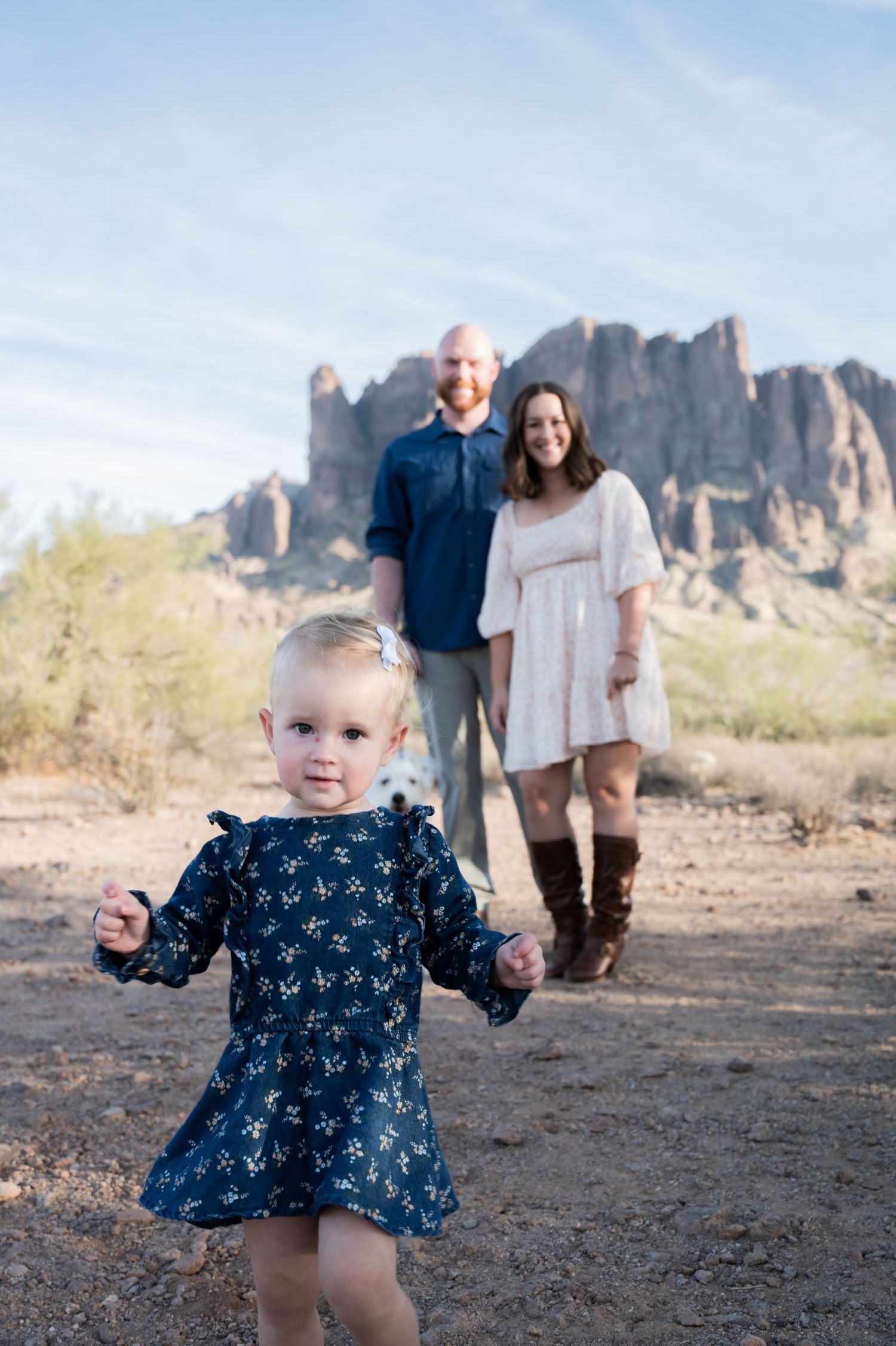 A toddler girl in a navy floral dress stands in front of her parents in an Arizona desert landscape with rocky mountains.