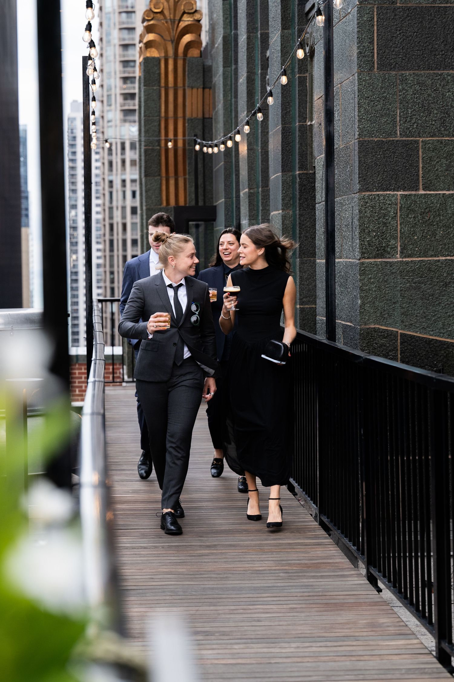 Wedding guests in formal attire walking along an urban rooftop balcony with string lights at The Pendry Hotel in Chicago Illinois.