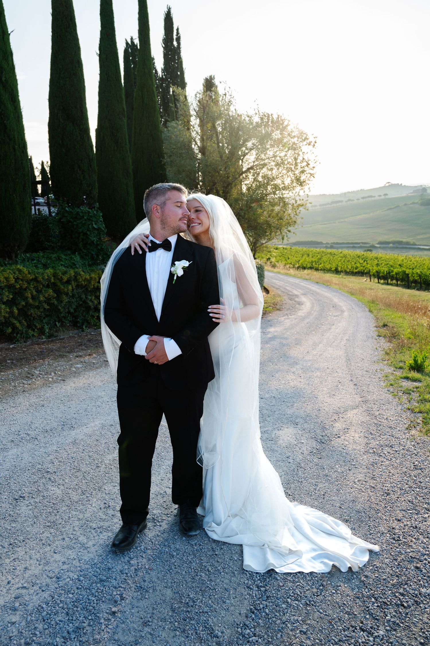 Happy bride and groom posing on a gravel path lined with cypress trees at sunset in Tuscany Italy.