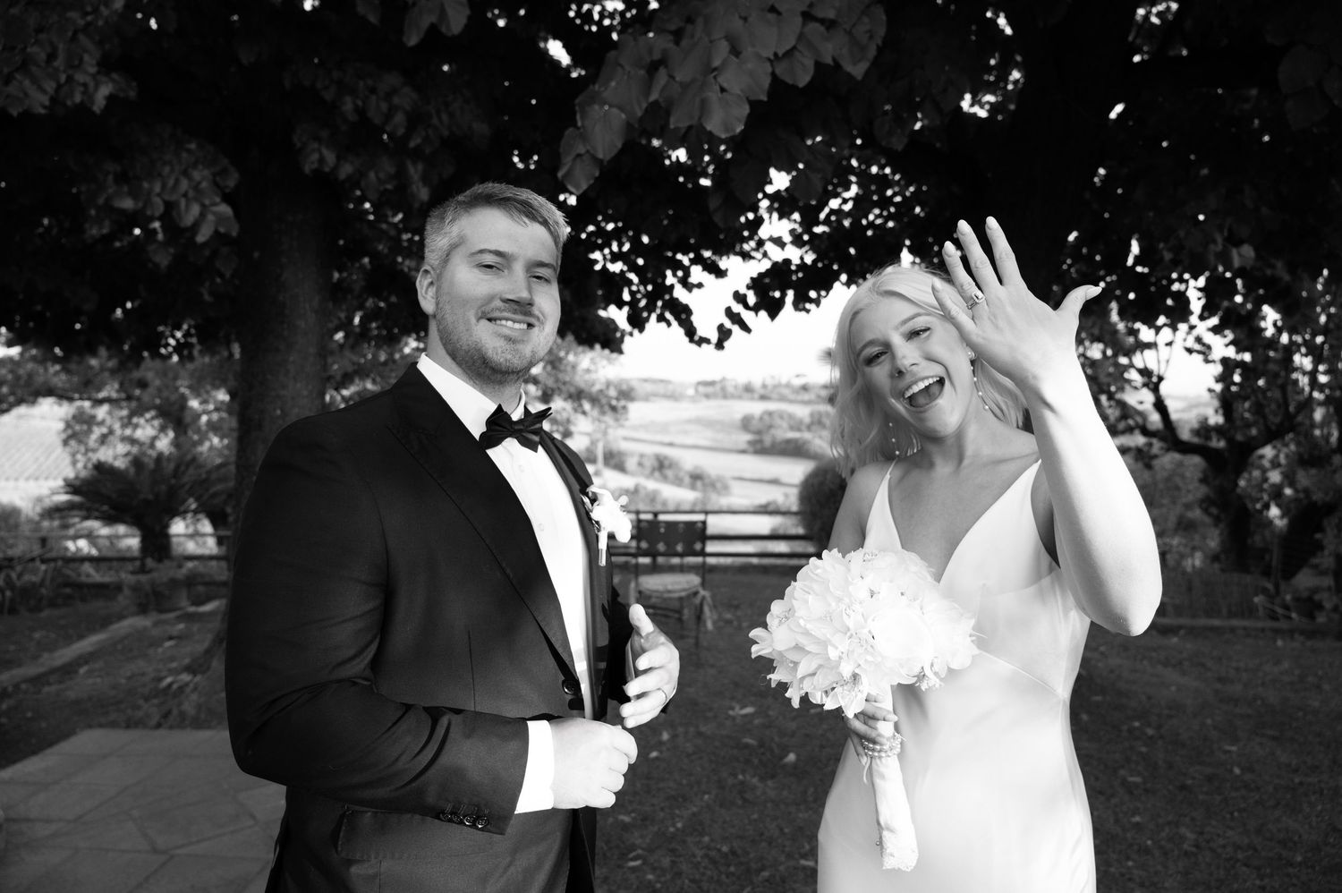 Black and white photo of smiling bride holding bouquet and groom in tuxedo posing outdoors under a tree.
