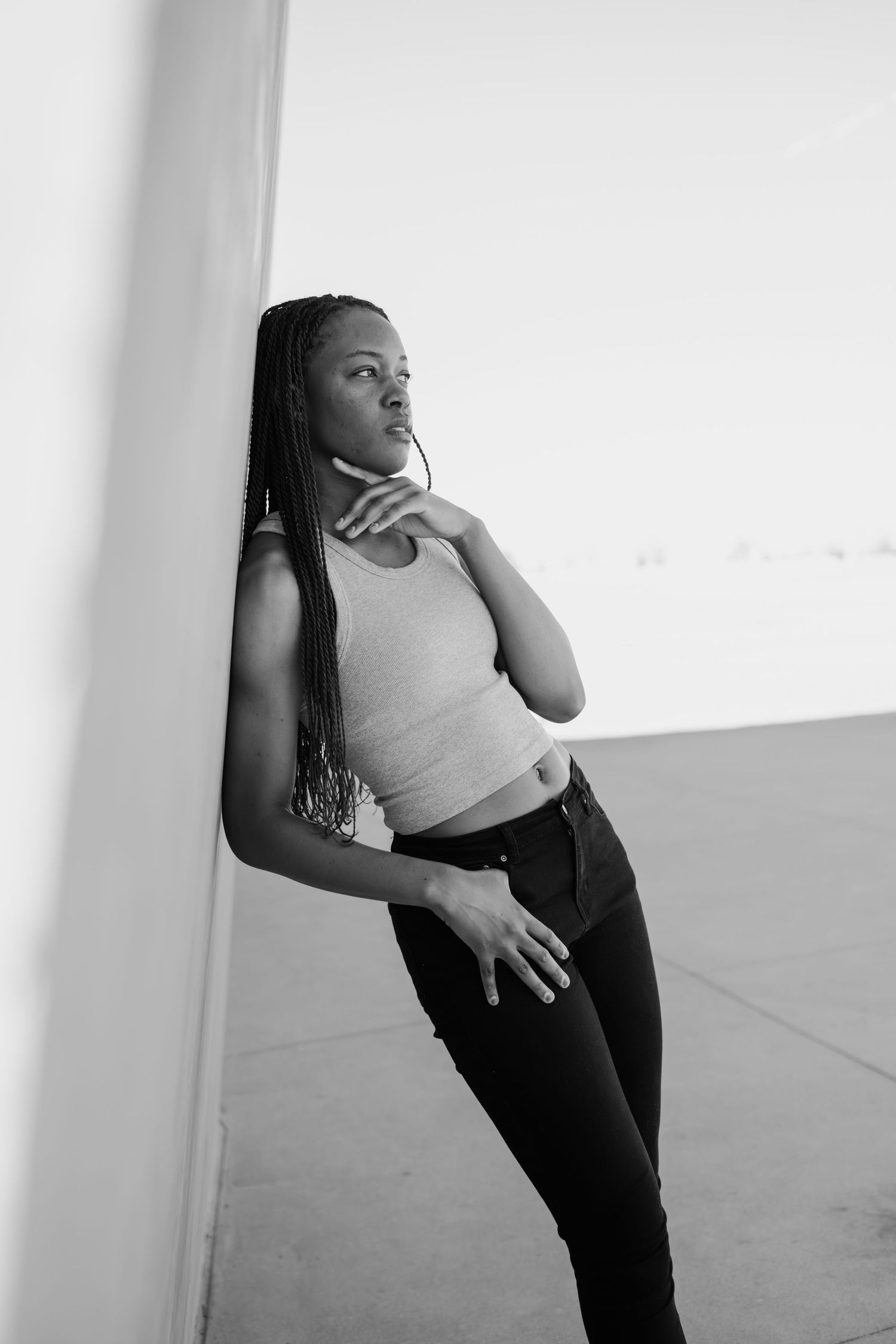 Black and white photo of a woman with braids leaning against a wall in thoughtful pose.