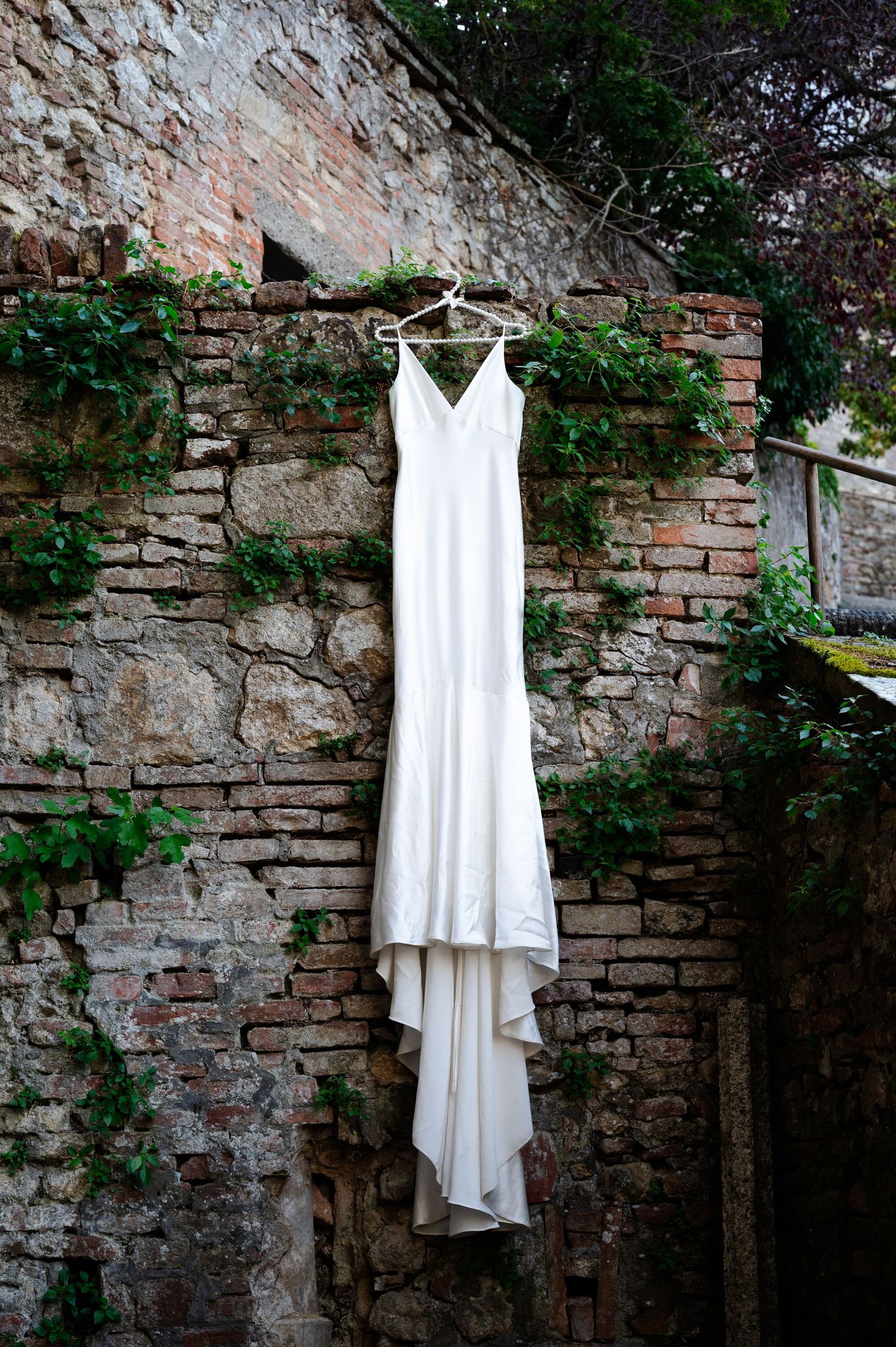 Elegant white satin wedding dress hanging on a rustic ivy-covered brick wall in Tuscany in Tuscany Italy.