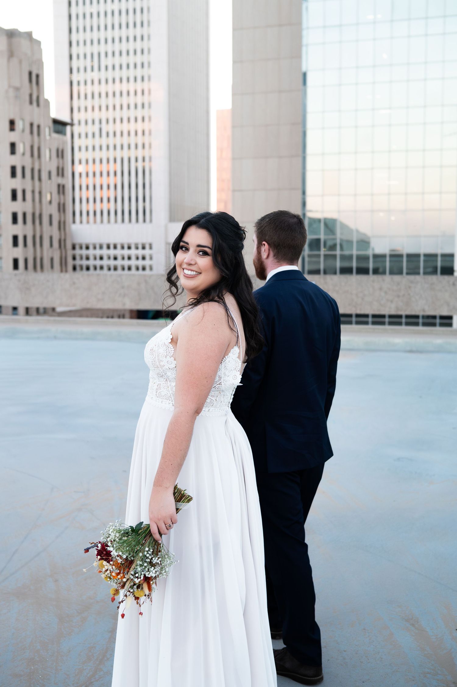 Bride in white gown holding colorful bouquet poses with groom on urban rooftop at sunset rooftop in phoenix