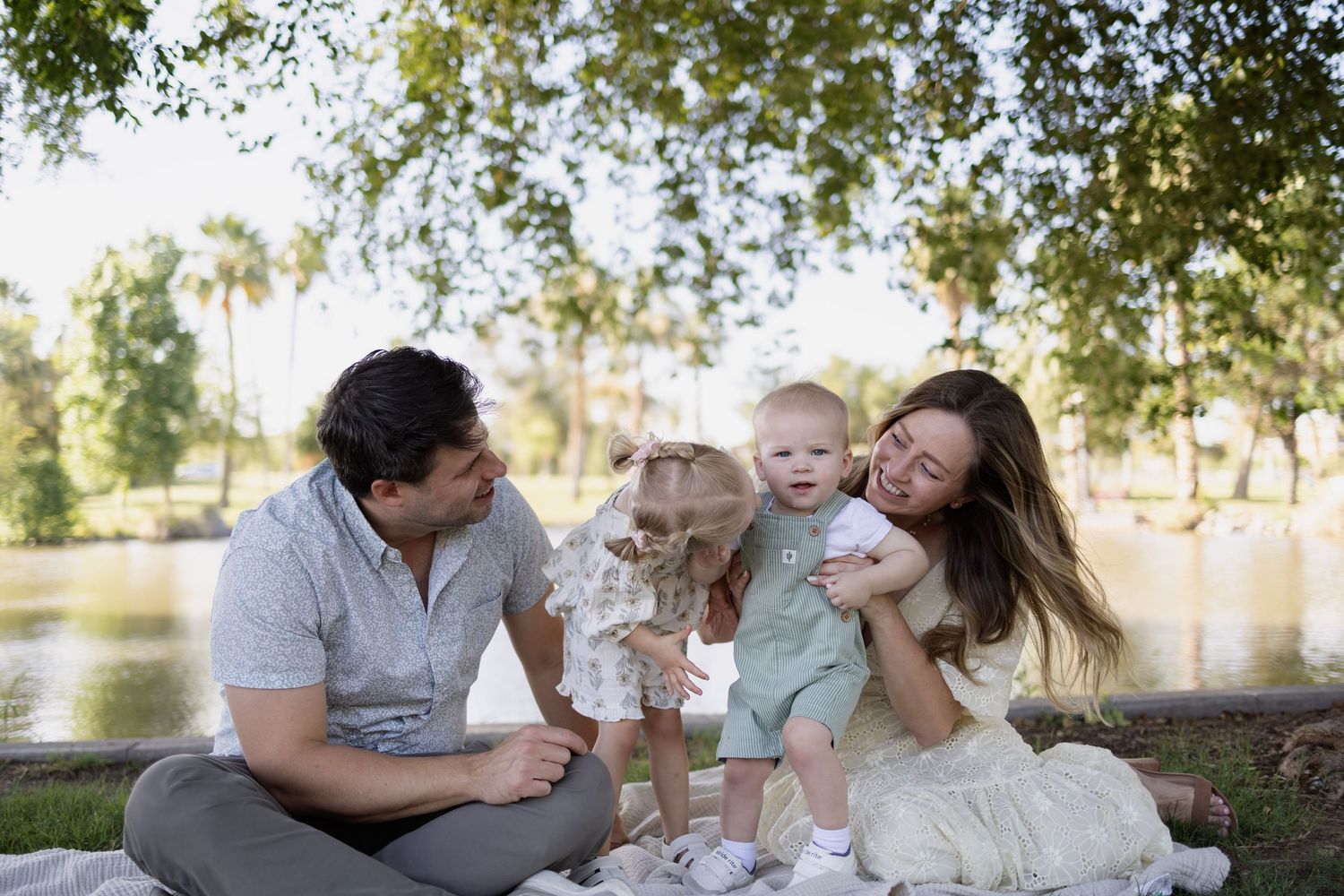Family of four enjoying a sunny day at the park by a lake, with parents smiling at their baby and toddler.