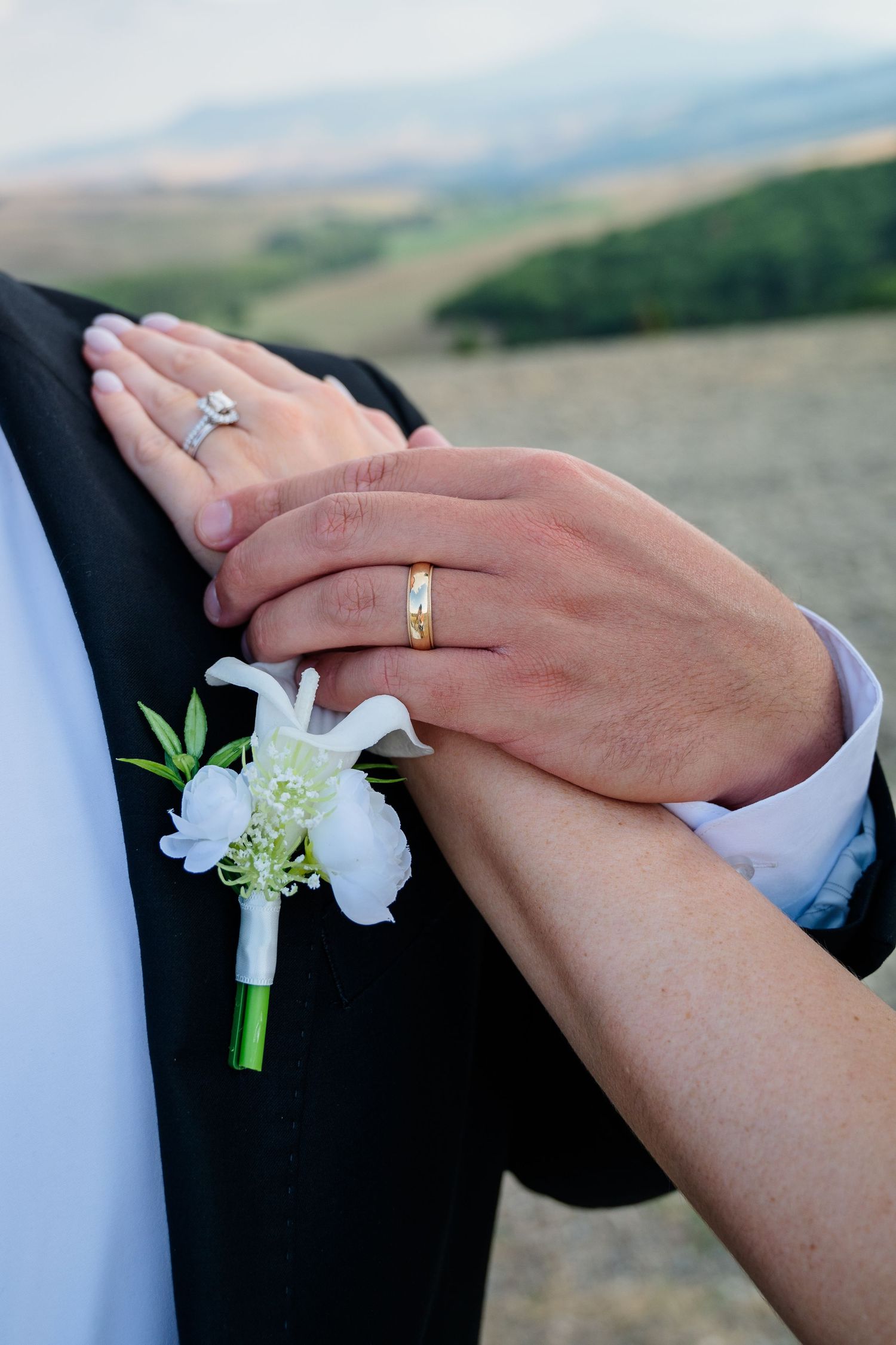 Close-up of bride and groom's hands showing wedding rings with white boutonniere against scenic landscape in Tuscany Italy.