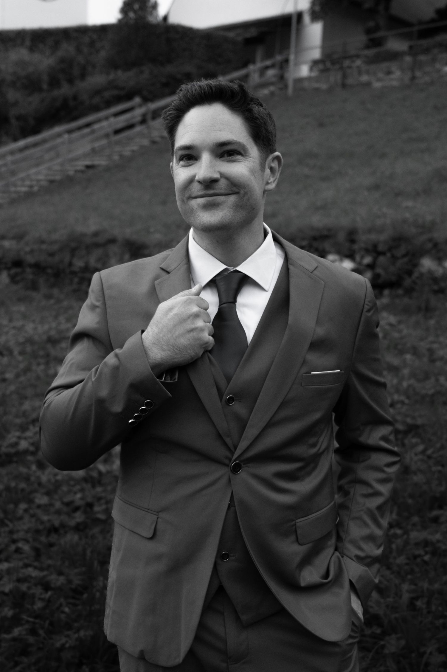 Dapper groom in three-piece brown suit adjusting tie on Swiss alpine hillside in black and white photo in Switzerland.