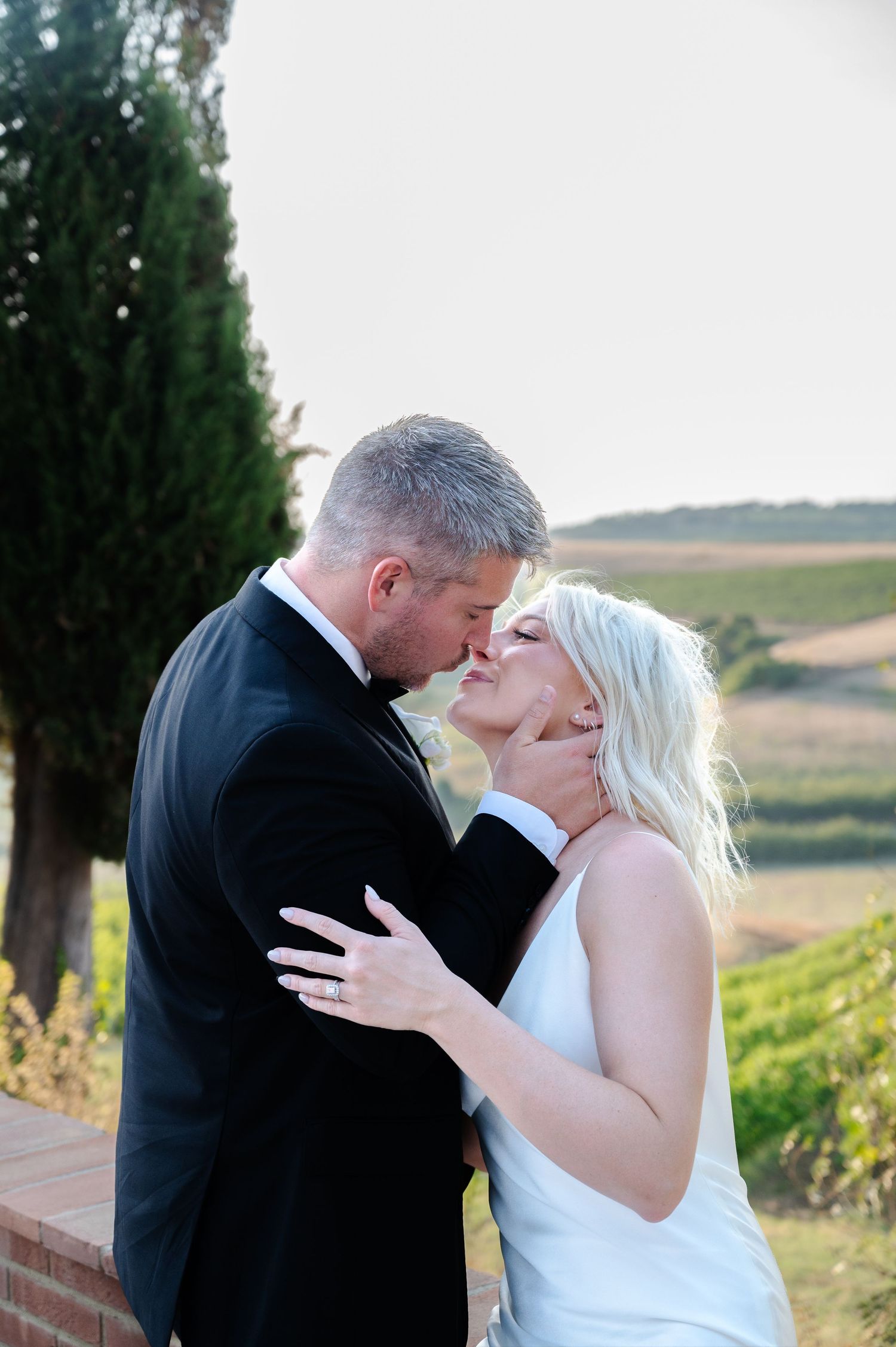 A bride and groom share a romantic kiss outdoors at a vineyard wedding venue on a sunny day.