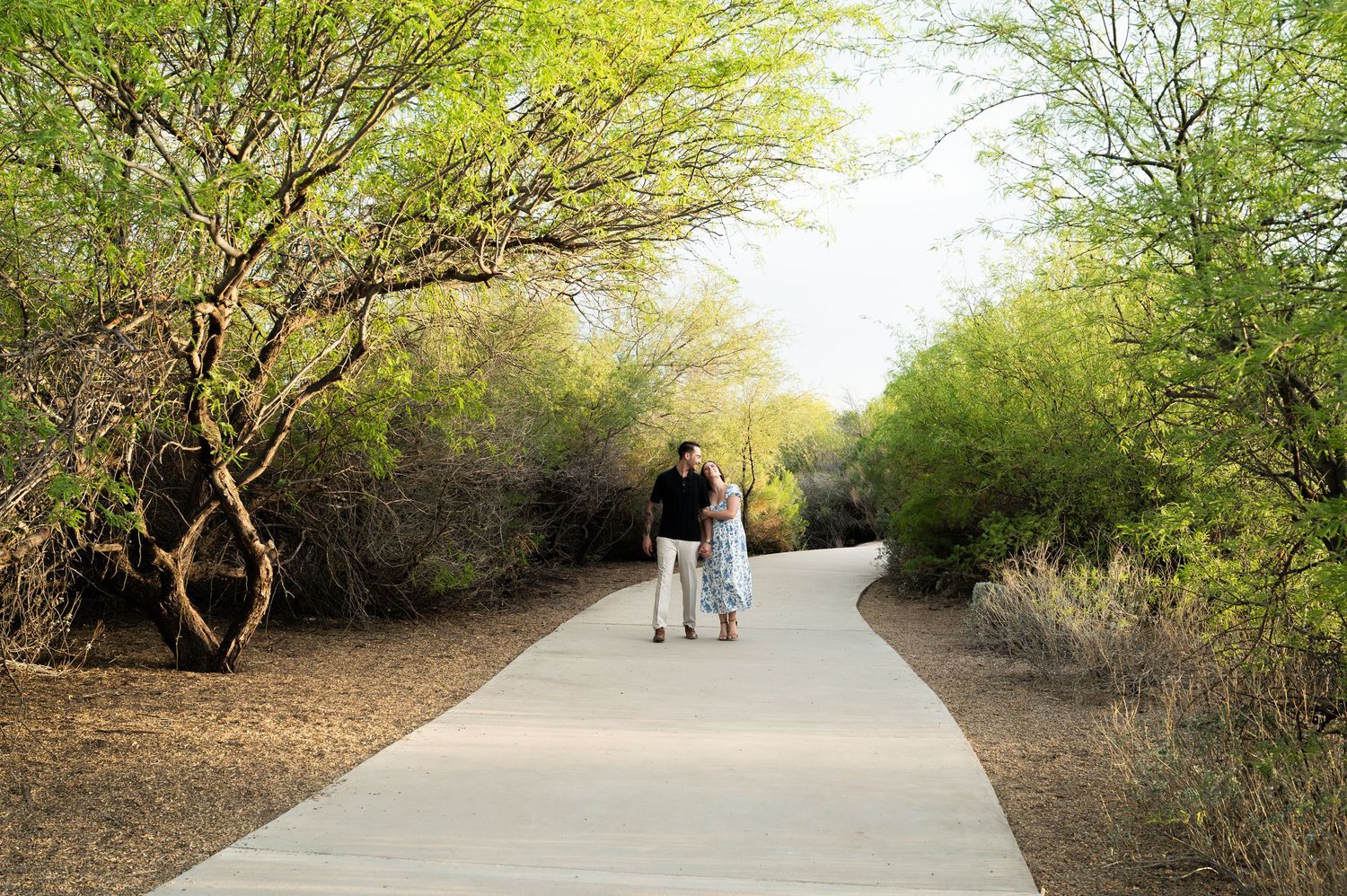 Couple walking hand-in-hand on a paved desert path surrounded by lush green trees and shrubs.