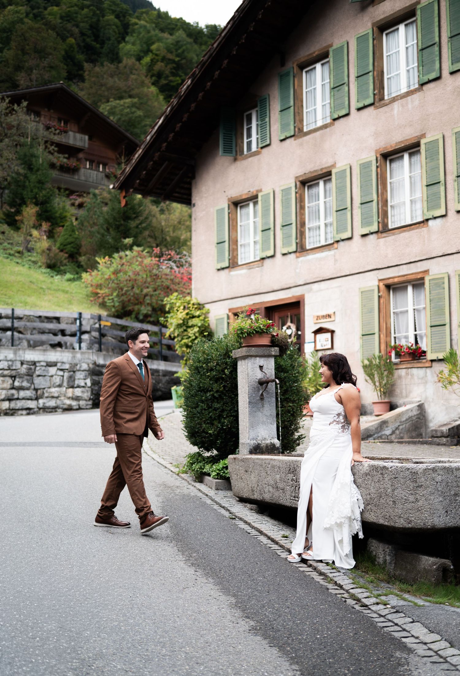 Bride and groom stroll past traditional Swiss chalet with green shutters on charming alpine village street in Lauterbrunnen Switzerland.