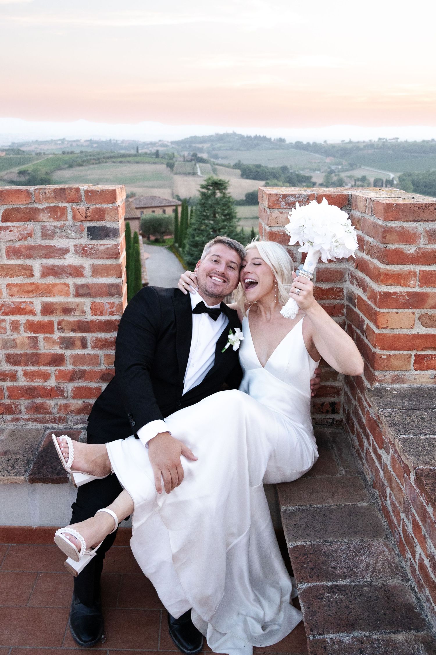 Joyful newlyweds sitting on brick terrace in Tuscany, bride in white gown holding bouquet, groom in tuxedo.