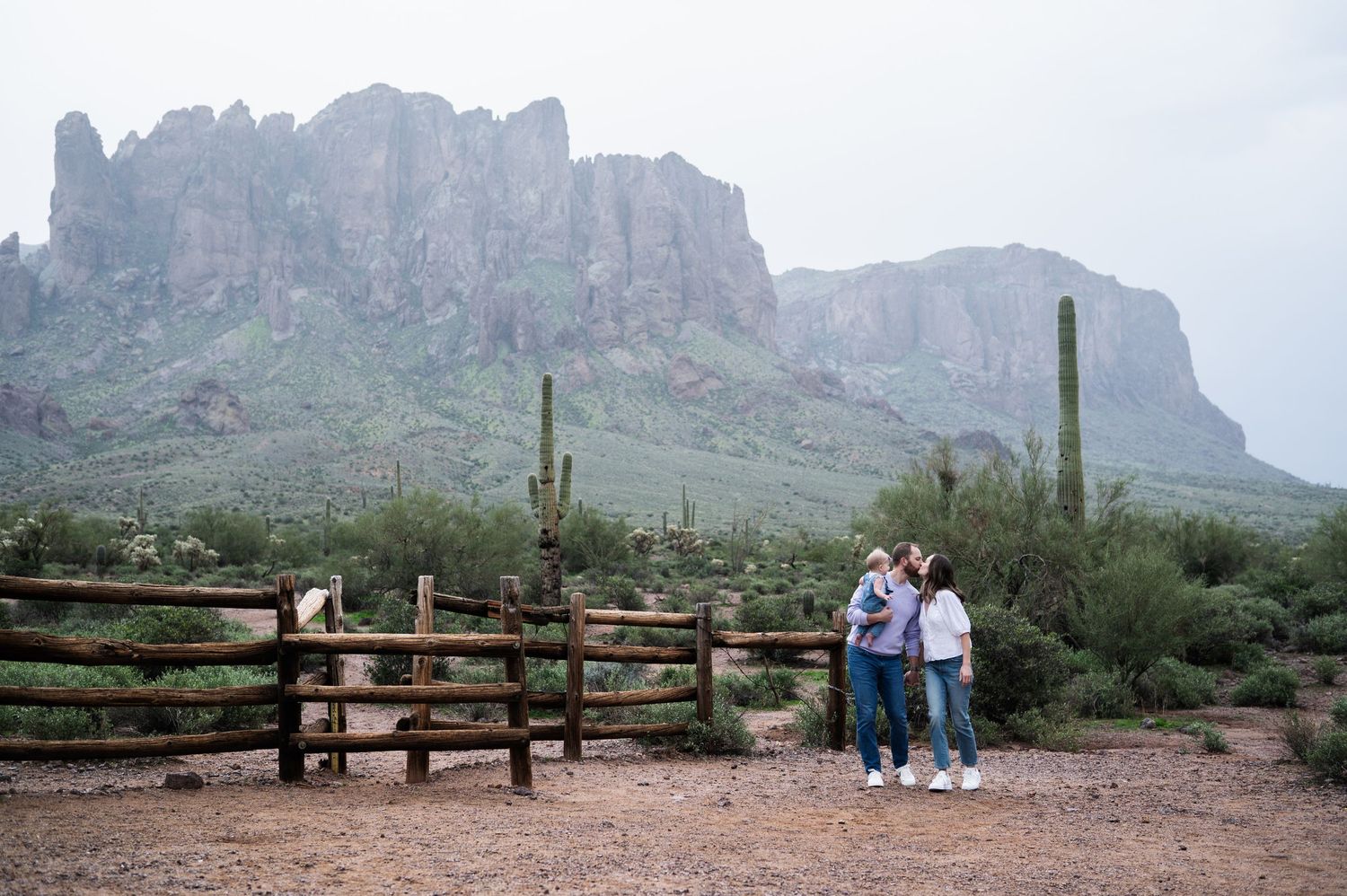 Two people stand by a rustic wooden fence with saguaro cacti and dramatic rocky Superstition Mountains in the Arizona desert.