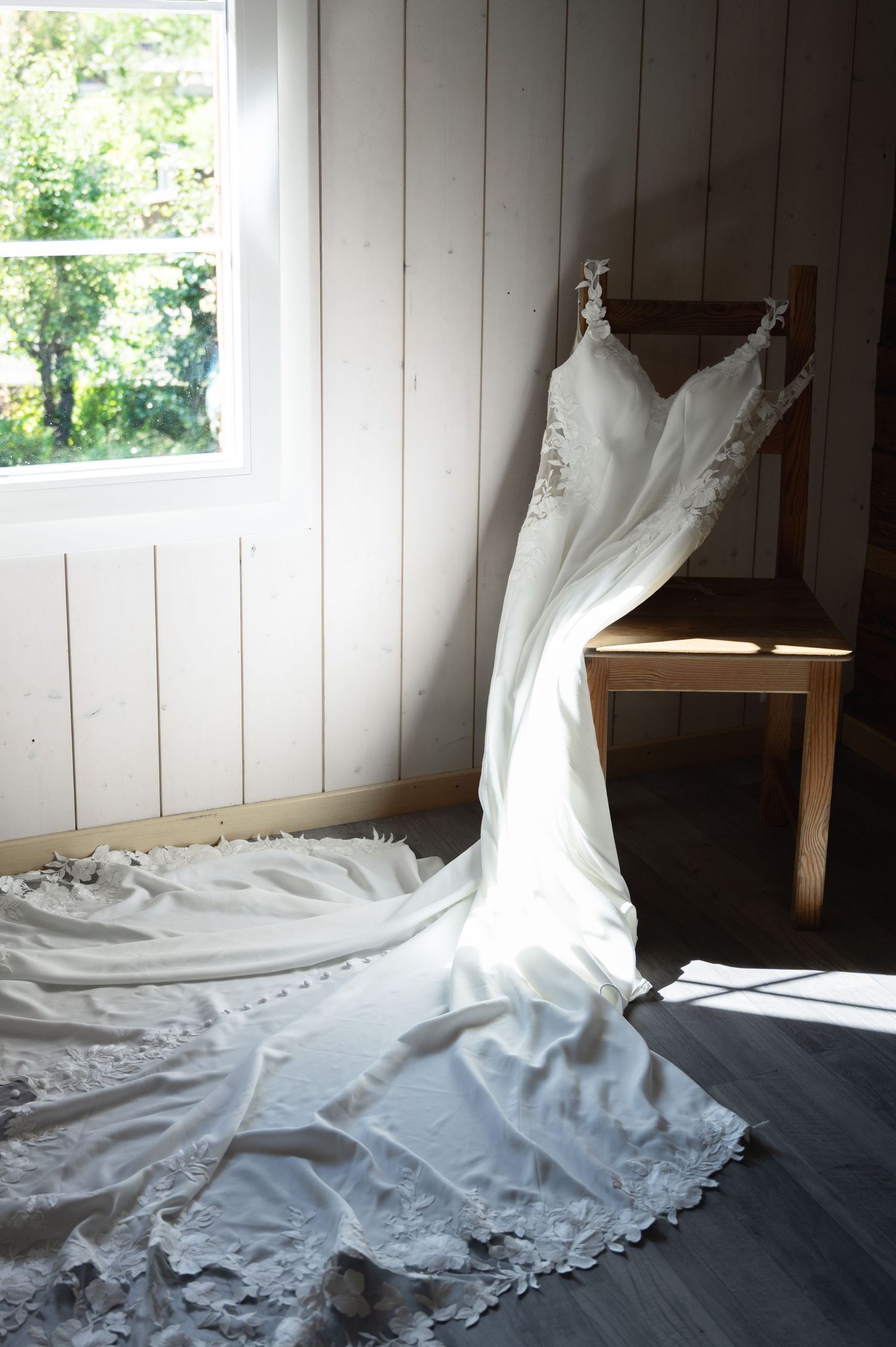 Elegant white lace wedding gown with long train draped over a wooden chair by a sunlit window in Lauterbrunnen Switzerland.