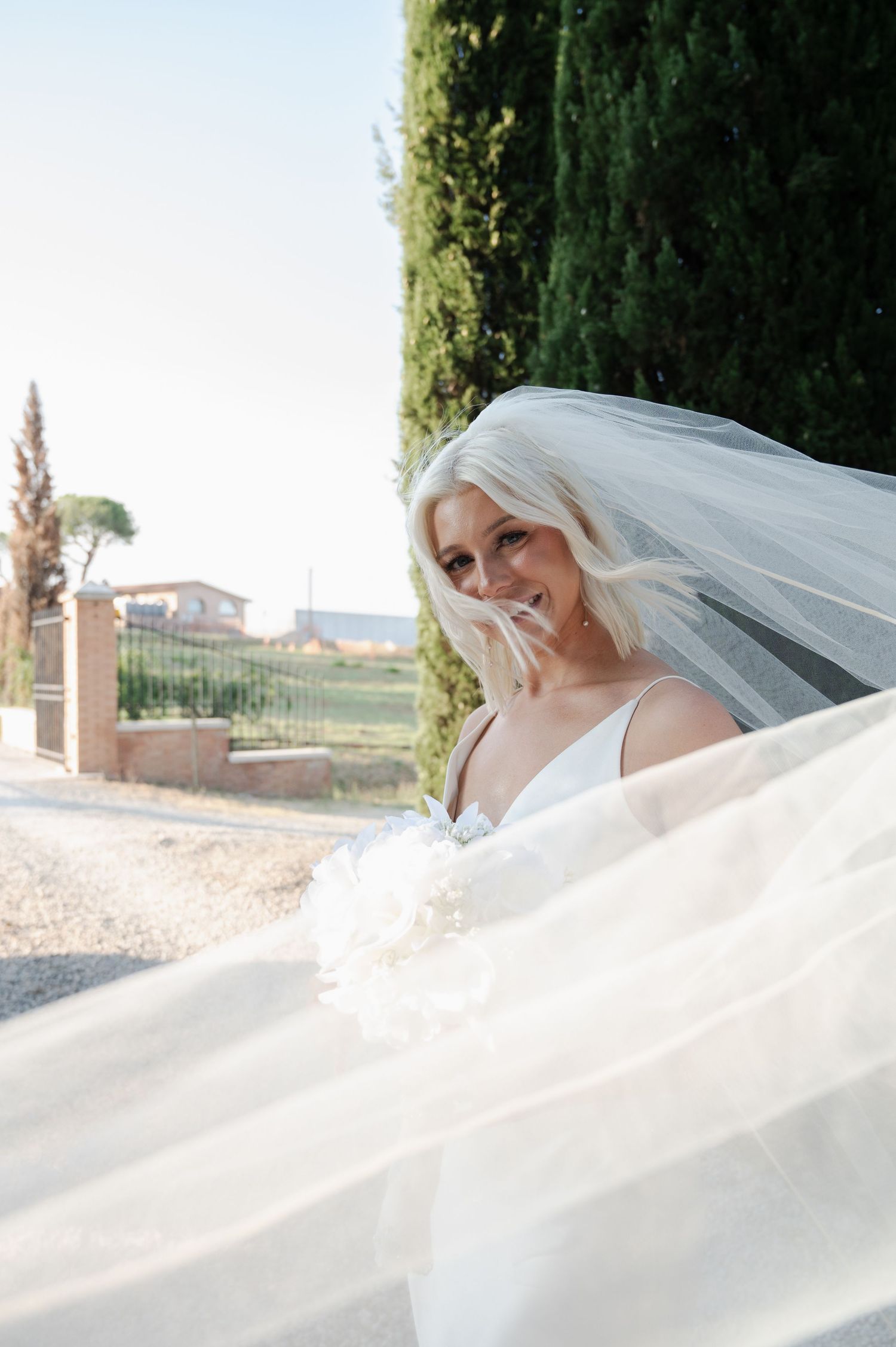 Radiant bride in white wedding dress with flowing veil billowing in breeze beside cypress trees in Tuscany Italy.