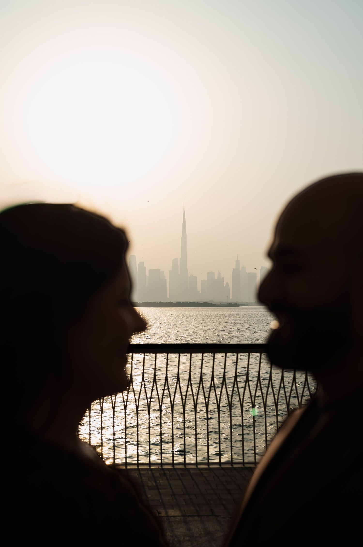 Silhouetted couple facing each other with Burj Khalifa and Dubai skyline across the water at sunset.