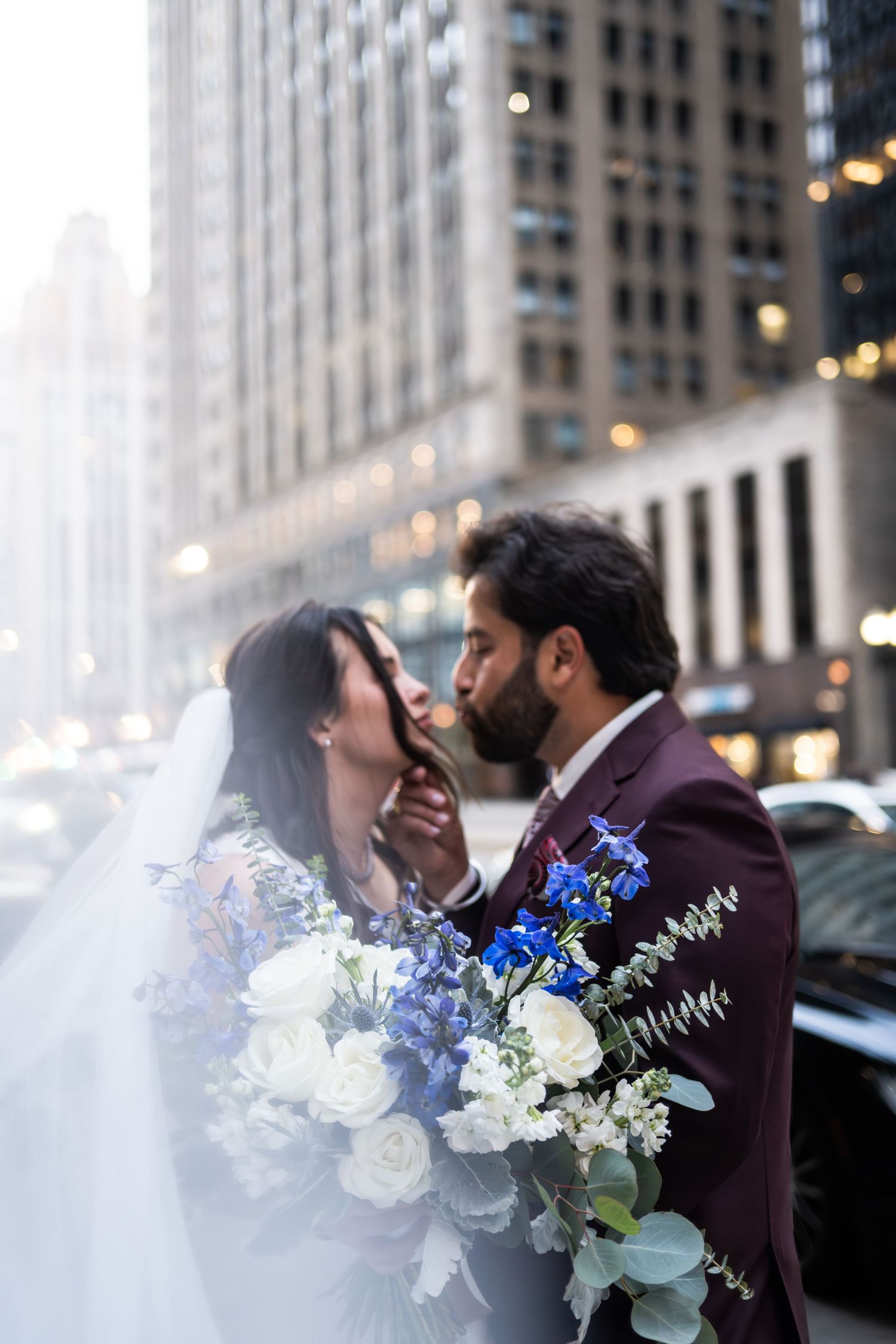 Groom in burgundy suit leans in to kiss bride holding blue and white floral bouquet on city street  at The Pendry Hotel in Chicago Illinois.