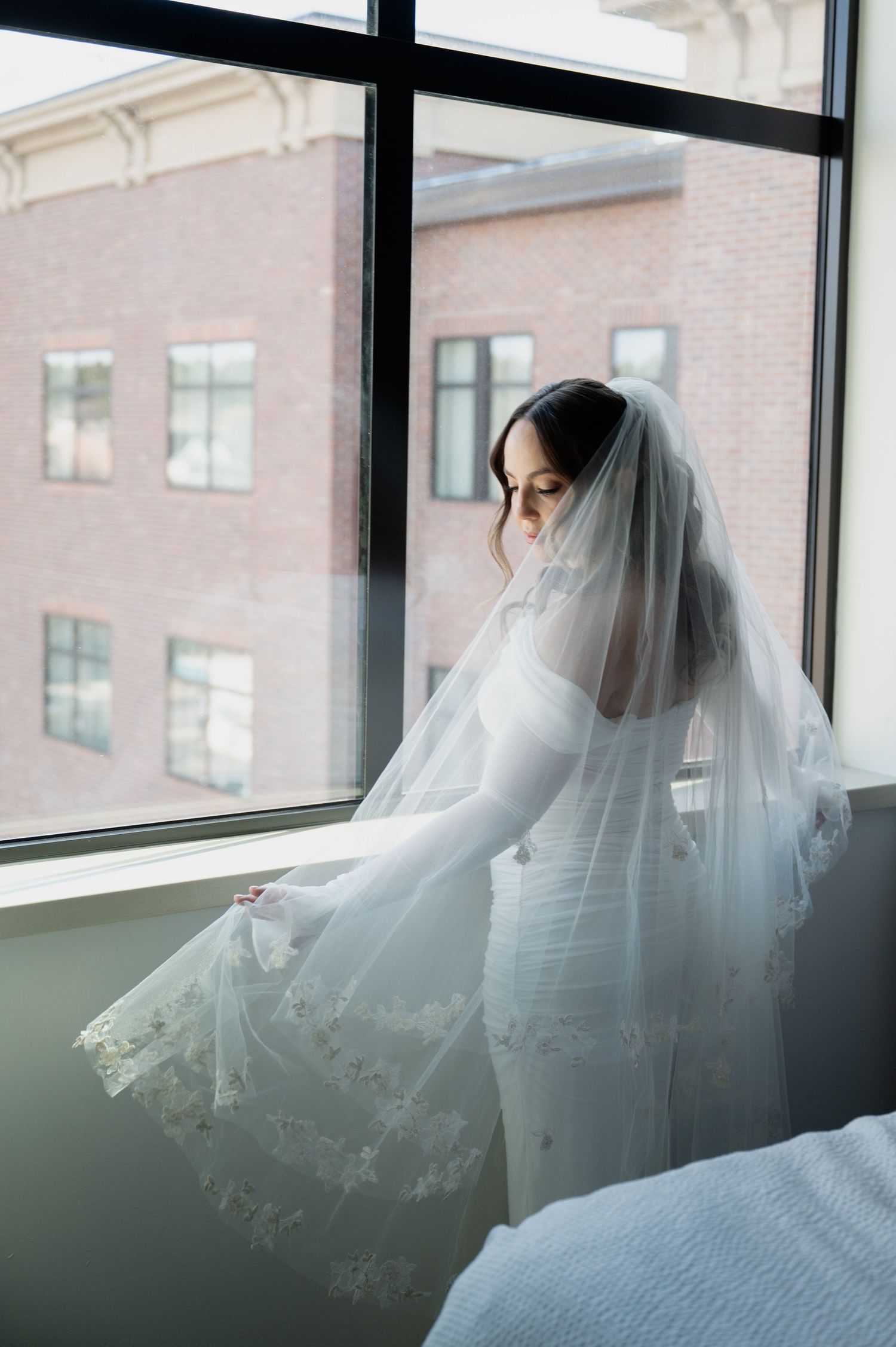 Bride in fitted white lace wedding gown and cathedral veil gazing out large window at brick building in Flagstaff Arizona.