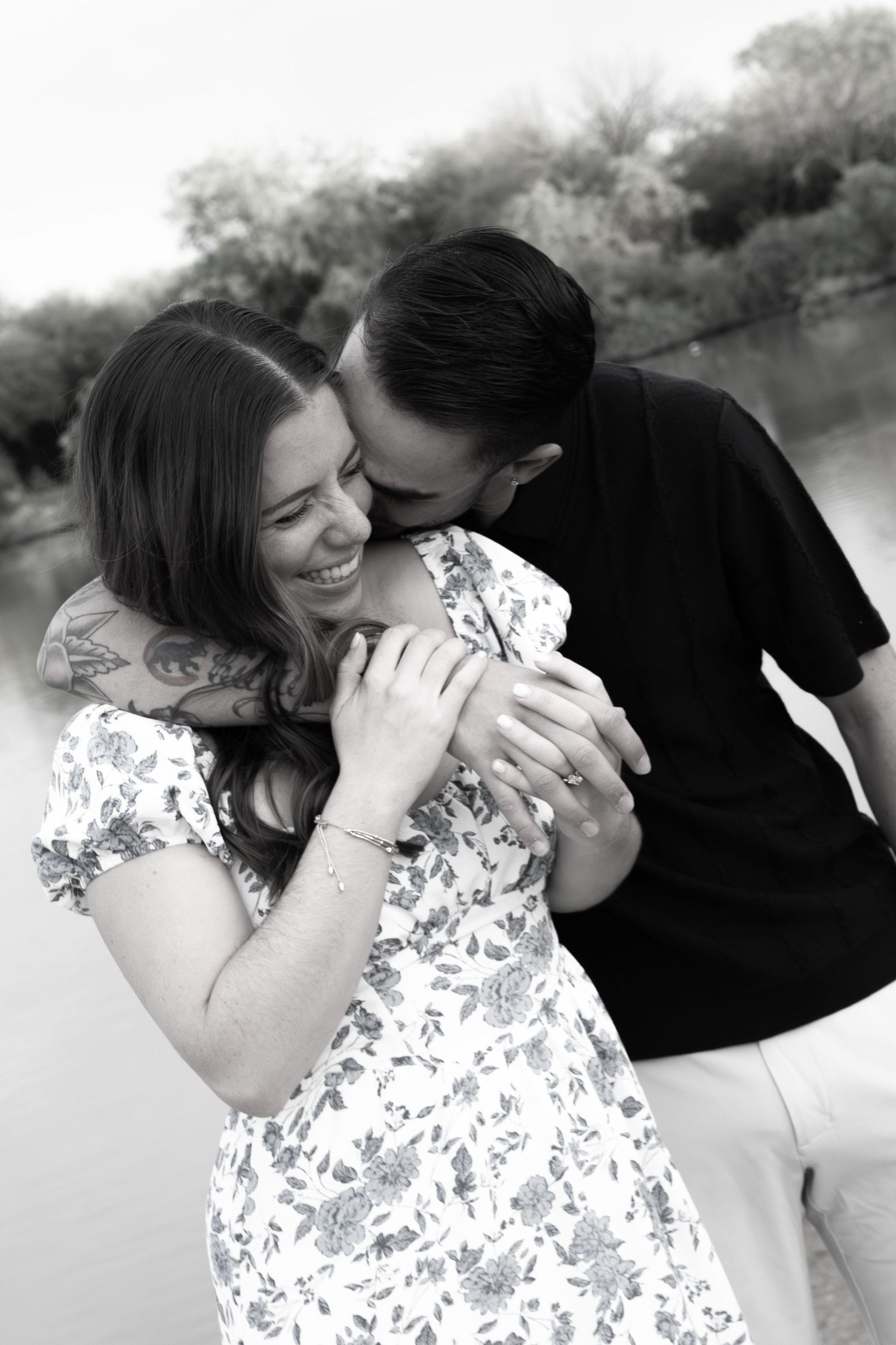 Black and white photo of a couple hugging and kissing on the cheek as she laughs joyfully near a lake in gilbert arizona for an engagement.