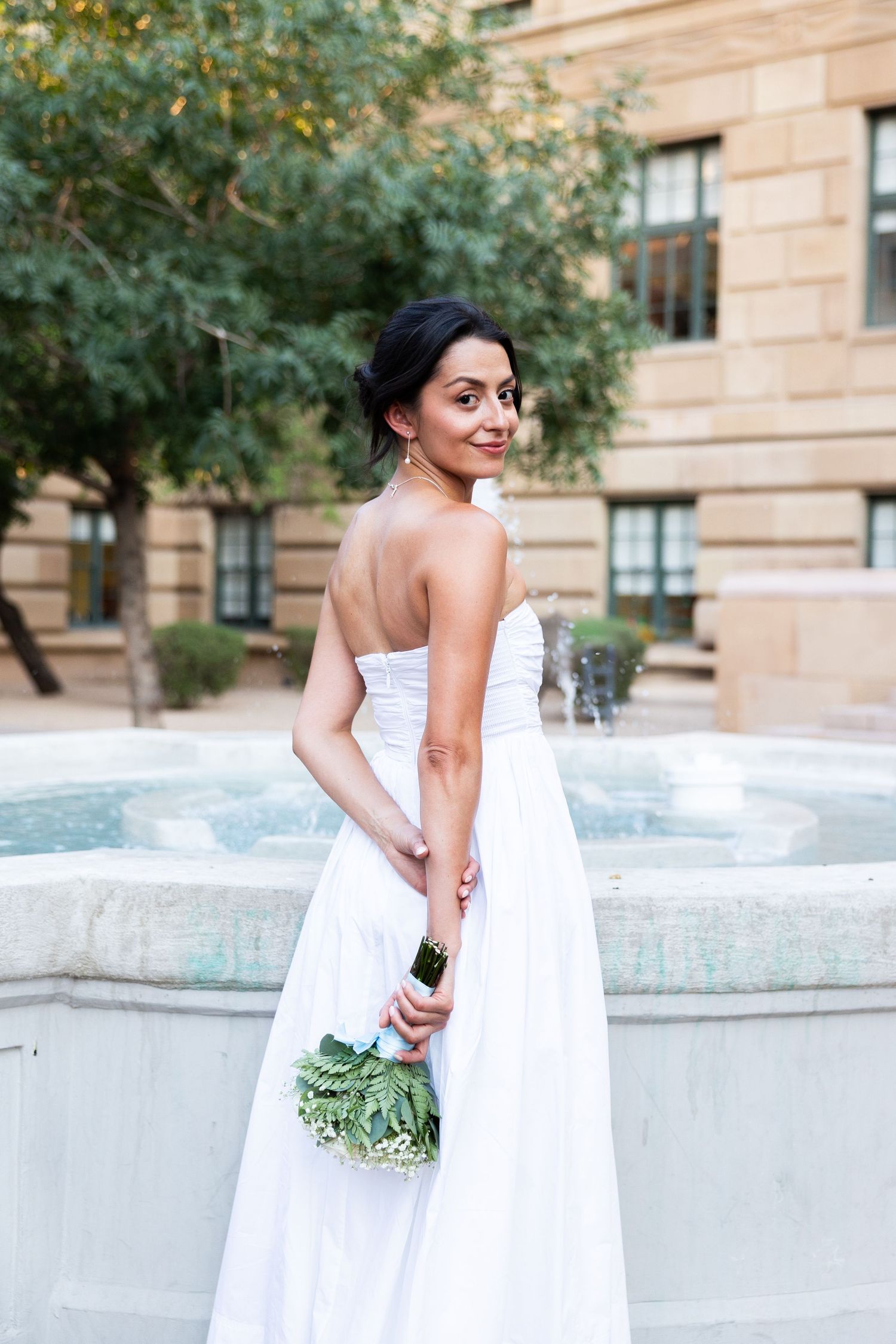 Elegant bride in strapless white gown holds bouquet near outdoor fountain at historic building at Old Courthouse in Phoenix Arizona.