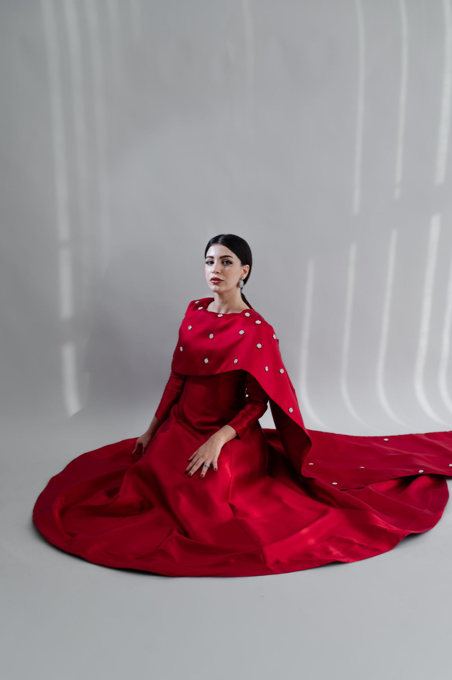 Woman in dramatic red gown with pearl-embellished cape poses gracefully in white studio setting in Jubail Saudi Arabia