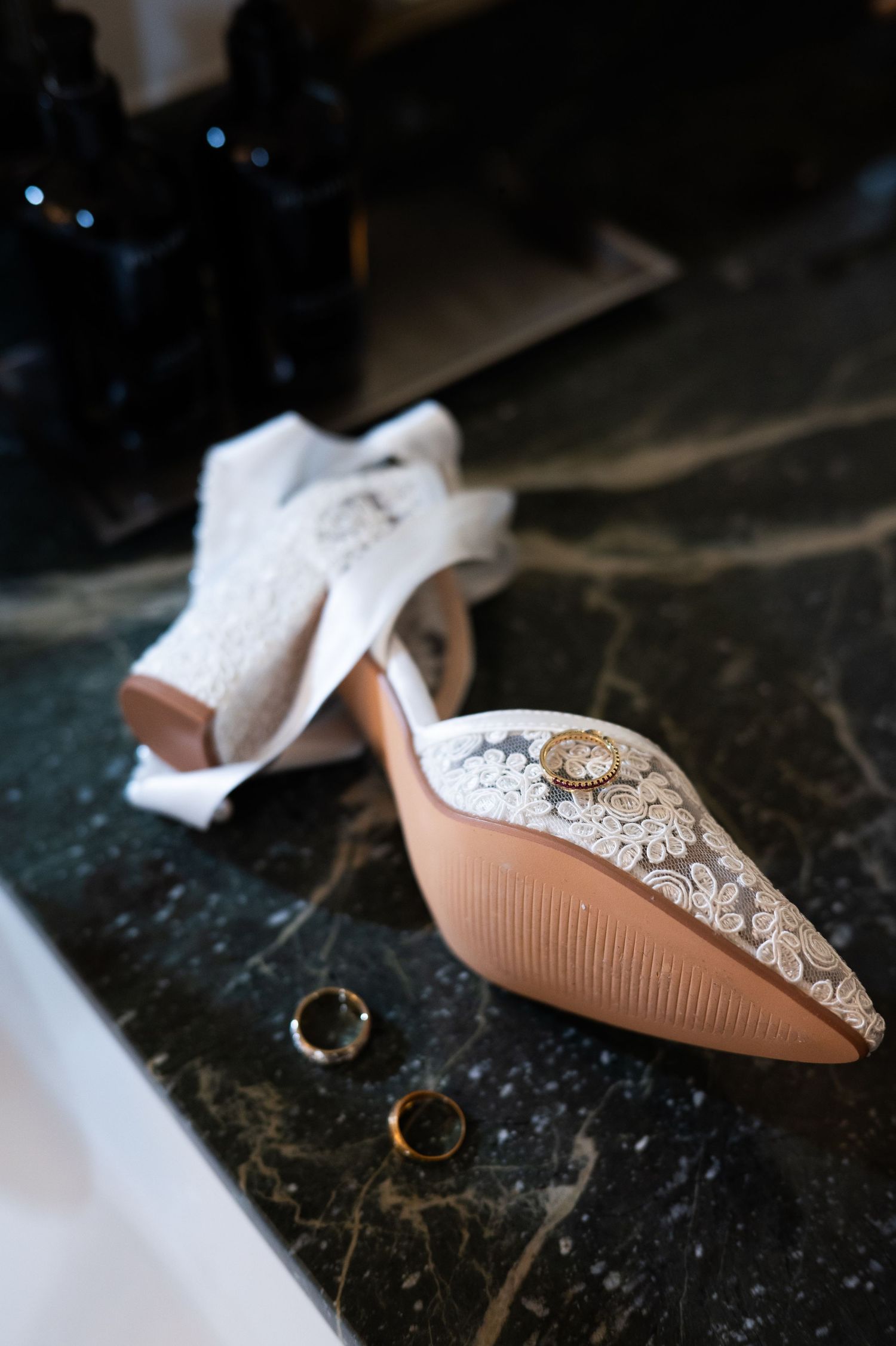 Lace bridal heels with jewel embellishment on marble surface beside gold wedding rings at The Pendry Hotel in Chicago Illinois.