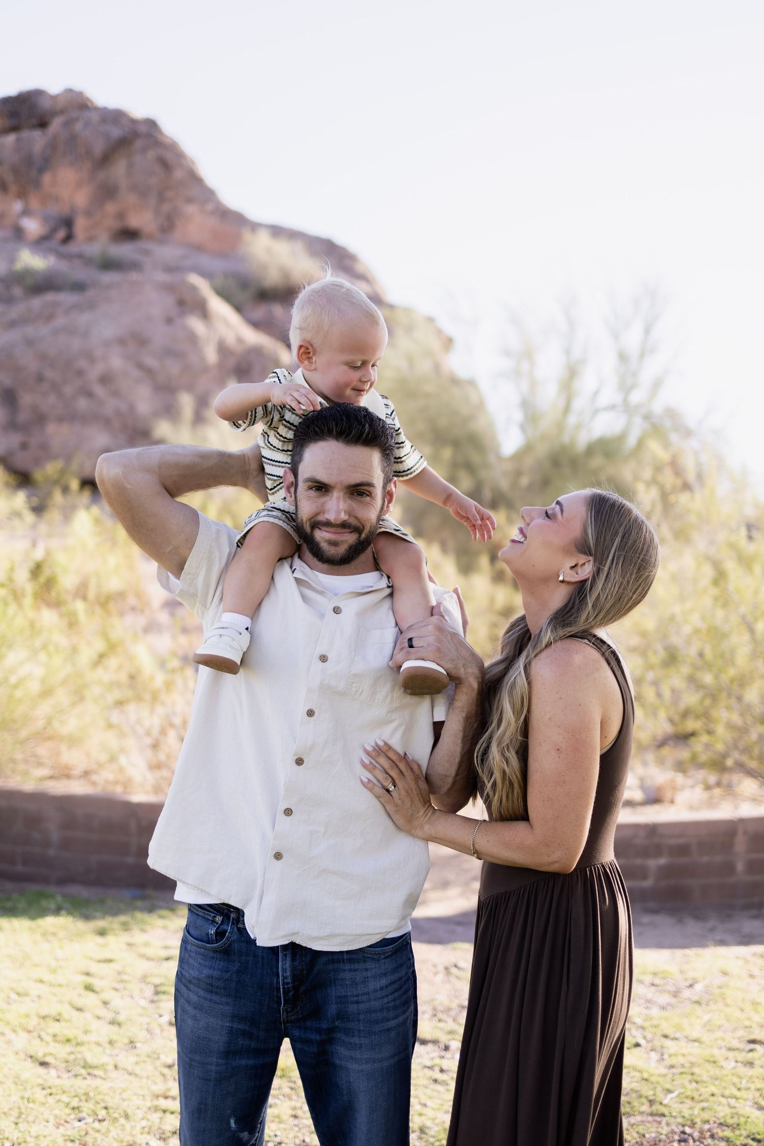 A happy young family poses outdoors, with dad carrying baby on shoulders while mom smiles, rocky desert landscape behind them.