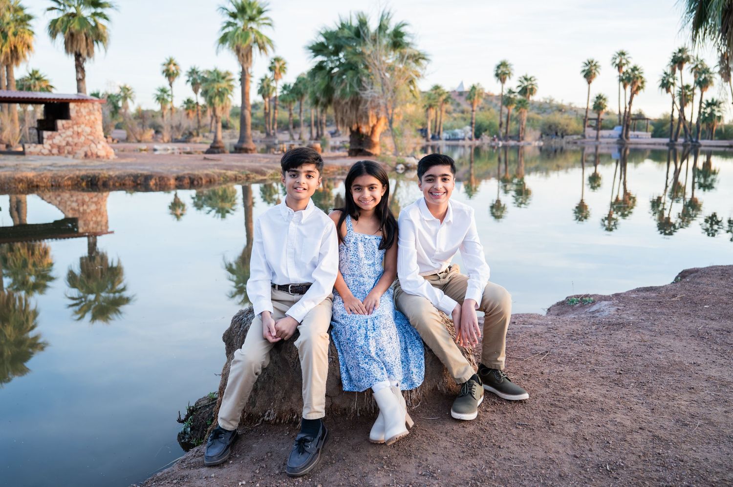 Three children sit on rocks by a reflective lake surrounded by palm trees in a warm outdoor setting.