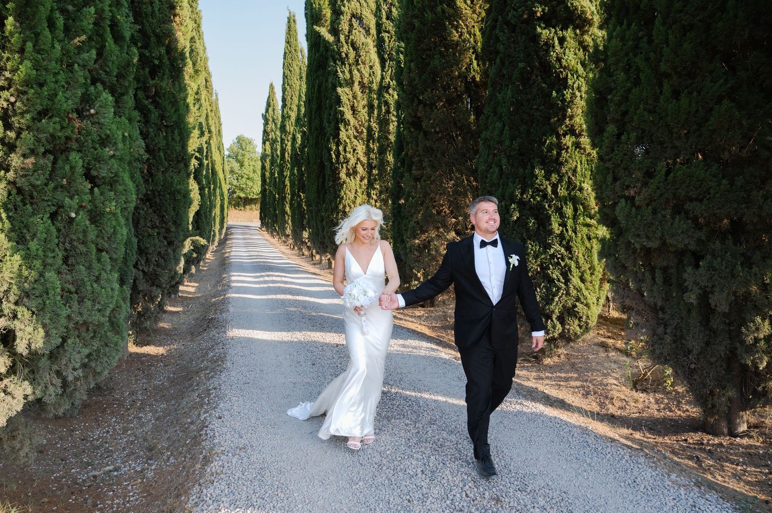 Newlyweds stroll together down a scenic cypress-lined driveway, bride in white gown and groom in black tuxedo in Tuscany Italy.