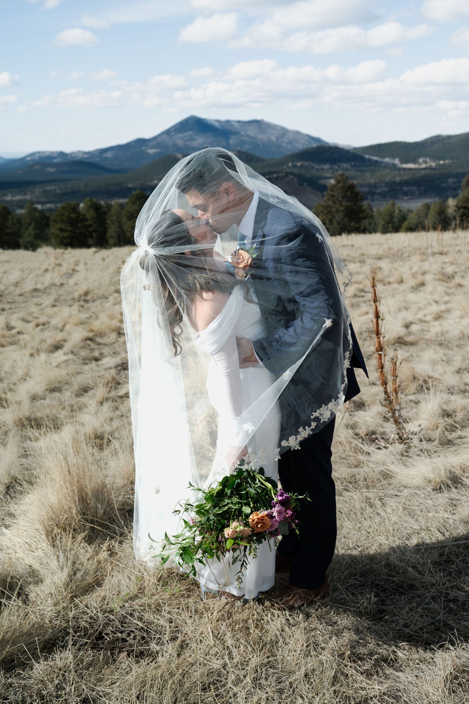 Bride and groom kiss under flowing veil with mountain landscape backdrop in Flagstaff in Flagstaff Arizona.