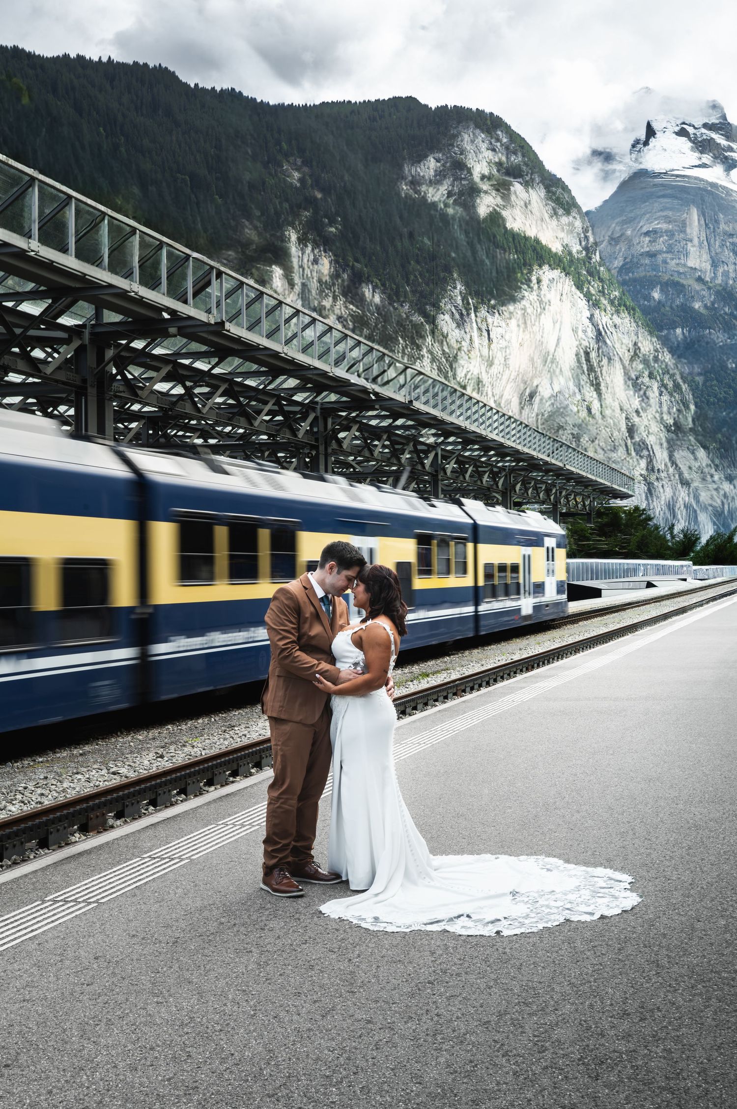 Bride and groom sharing a romantic kiss on a Swiss mountain train station platform with passing train.